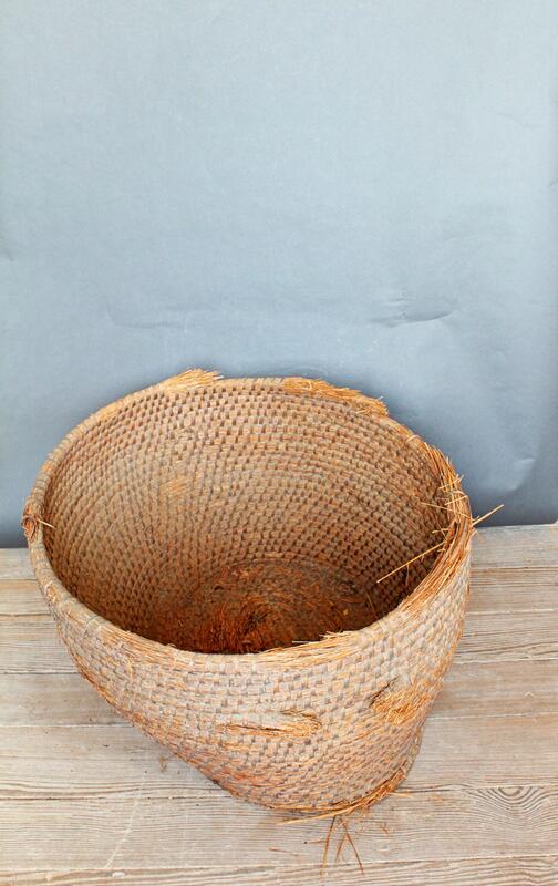 A large, round, woven basket with a slightly frayed edge sits on a wooden floor against a gray wall. The basket is empty and appears to be made from natural fibers.