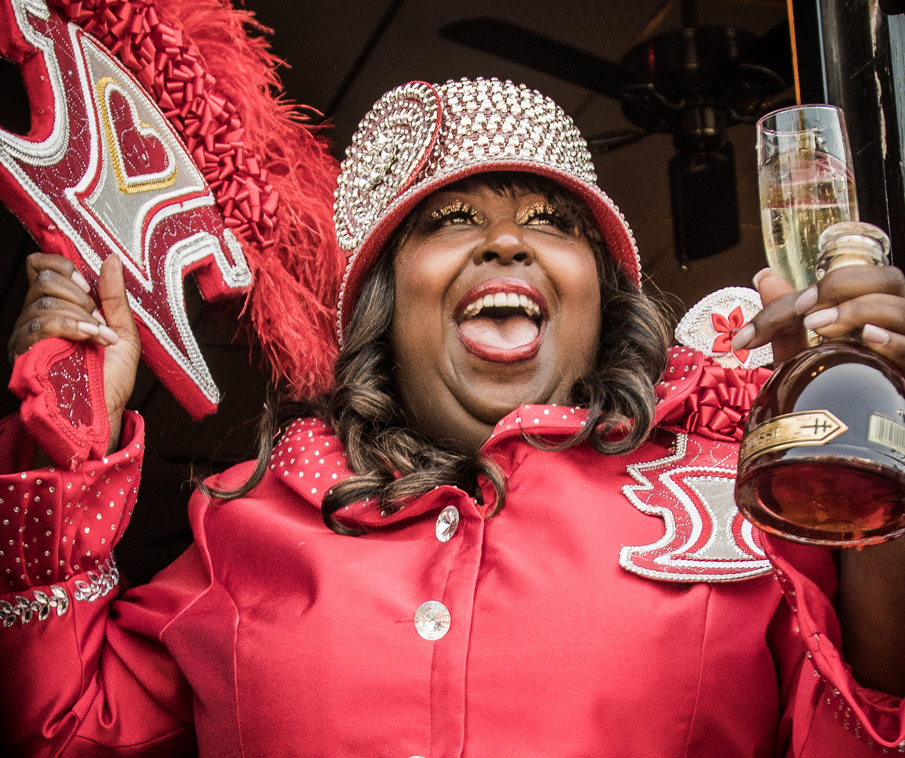 A person wearing a vibrant red outfit and a sequined hat joyfully holds a decorated fan and a glass of champagne. They are smiling broadly, exuding a celebratory and lively atmosphere.