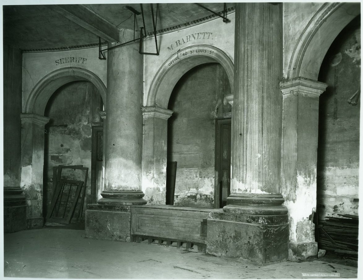 A black and white photo of an old, deteriorating interior with large pillars and arched doorways. The words Sheriff and W. Barnett are inscribed above the arches. The area shows signs of wear, with peeling paint and scattered debris.