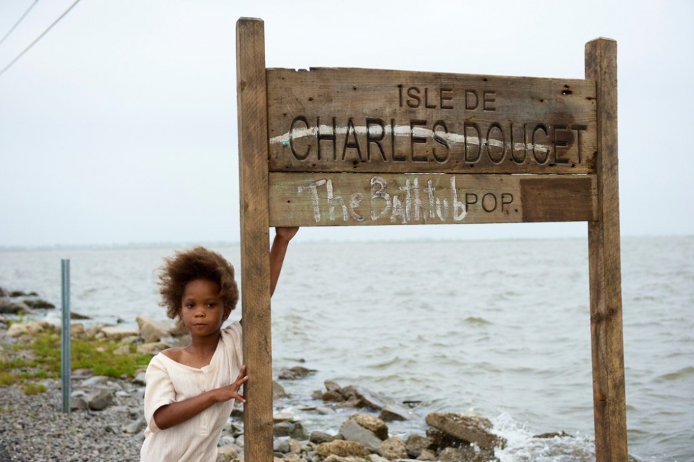 A young child with curly hair stands holding a wooden sign that reads Isle de Charles Doucet The Bathtub Pop. The sign is near a rocky shore with water visible in the background under an overcast sky.