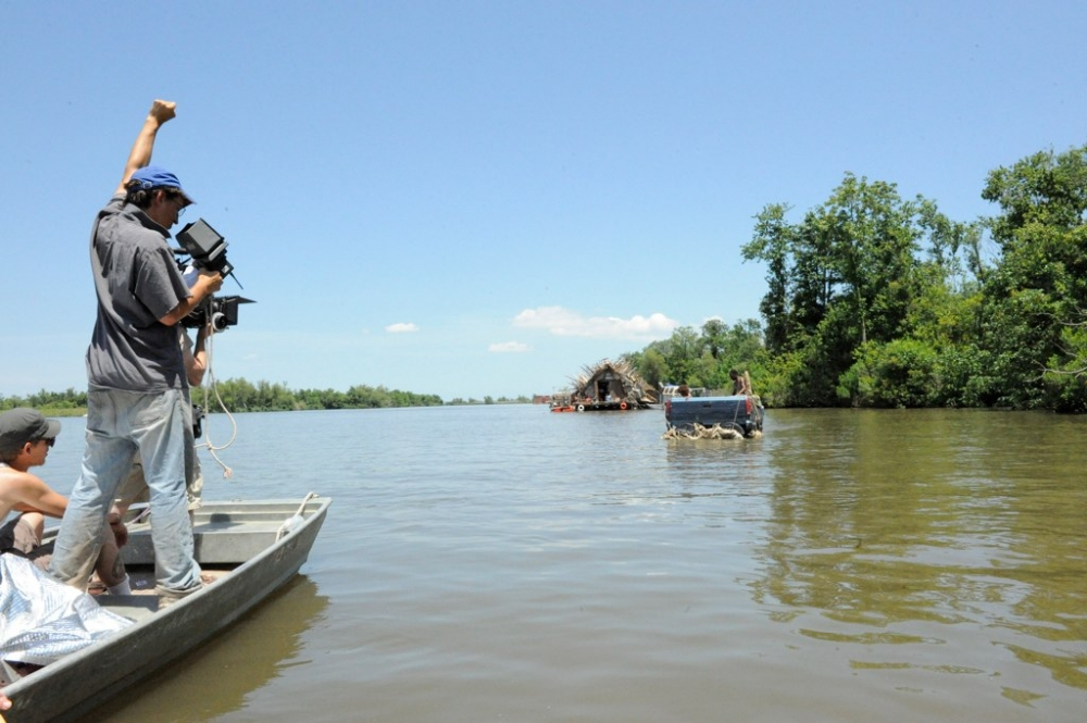 A filmmaker operates a camera on a boat in a wide river, capturing footage of a floating, rustic house surrounded by lush greenery under a clear blue sky. Another person steers the boat.
