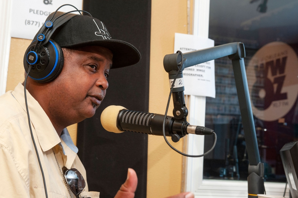 A person wearing a cap and headphones speaks into a radio microphone in a studio. The background features posters and a window showing a radio stations logo.