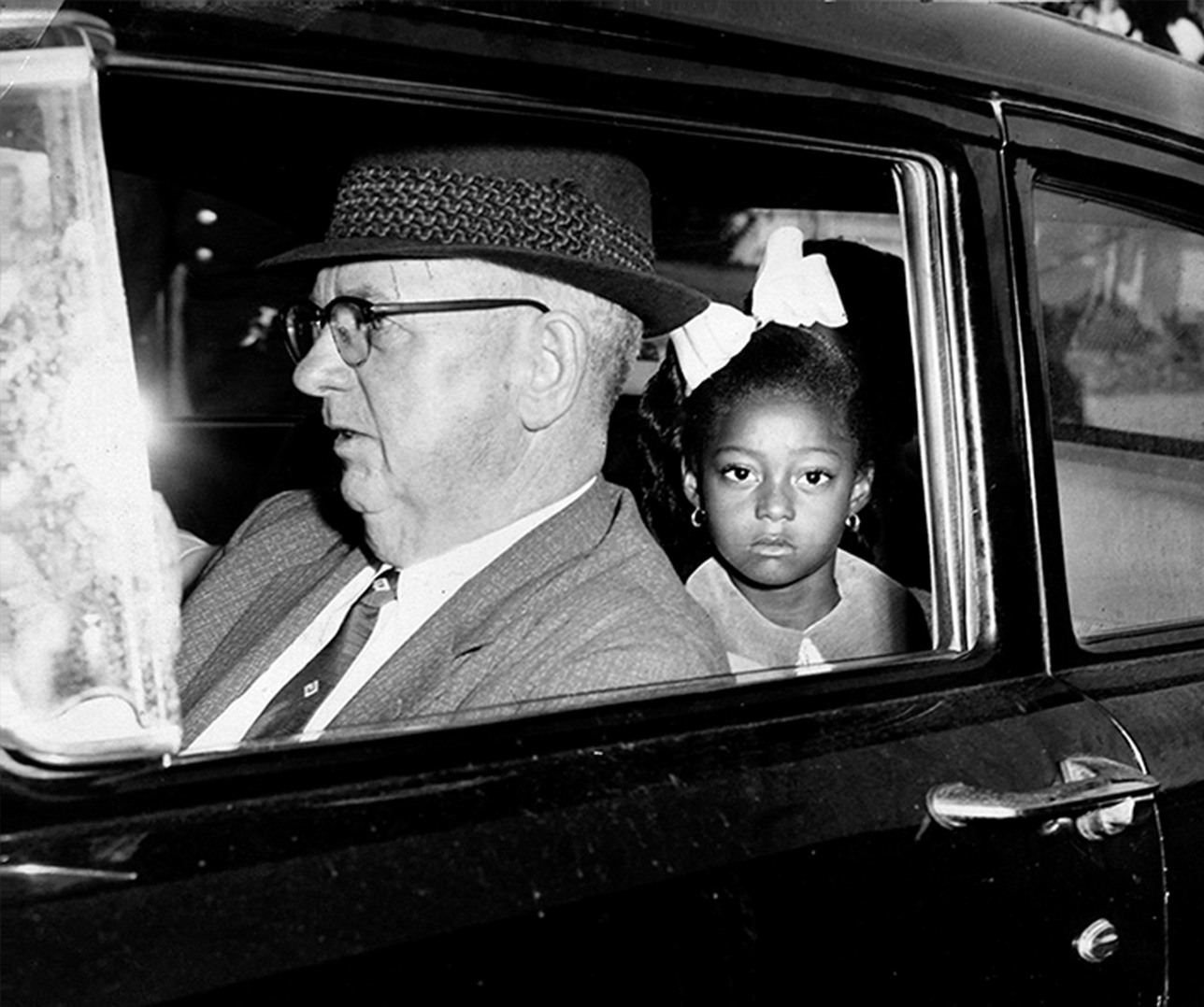 A young girl with a white bow in her hair sits in the backseat of a car, looking out the window. An older man wearing a hat and glasses is in the drivers seat, focused on the road. The car appears to be from the mid-20th century.