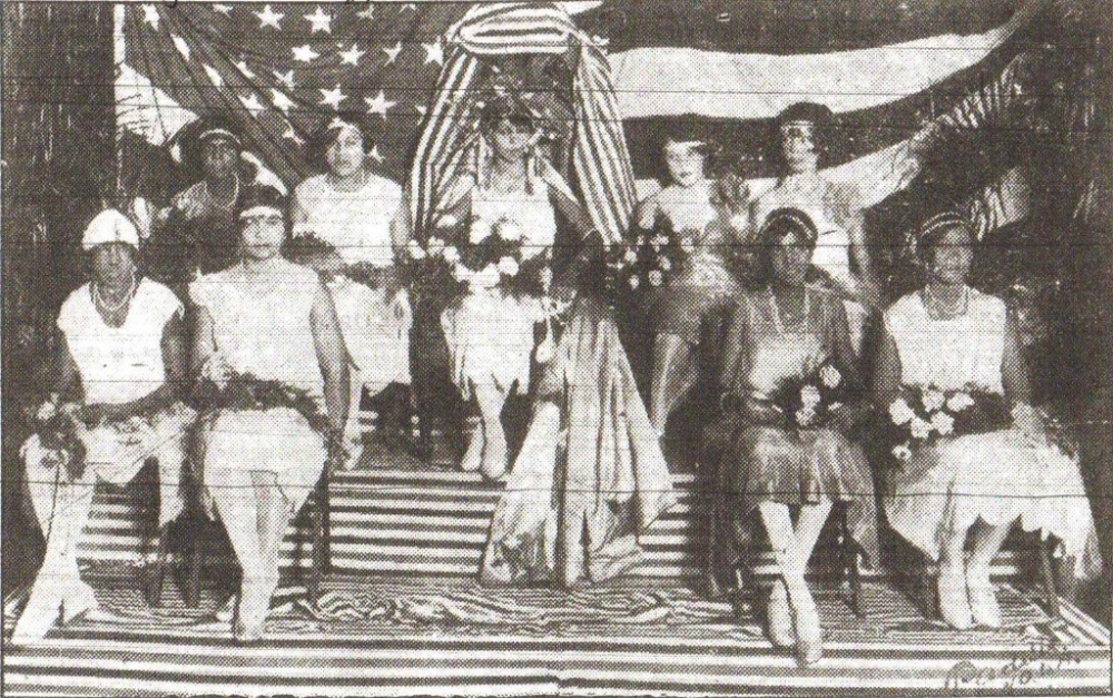 A vintage photo of a group of women seated and standing on a striped stage. They are dressed in formal attire and holding bouquets. An American flag serves as the background, and a large draped fabric hangs above them.