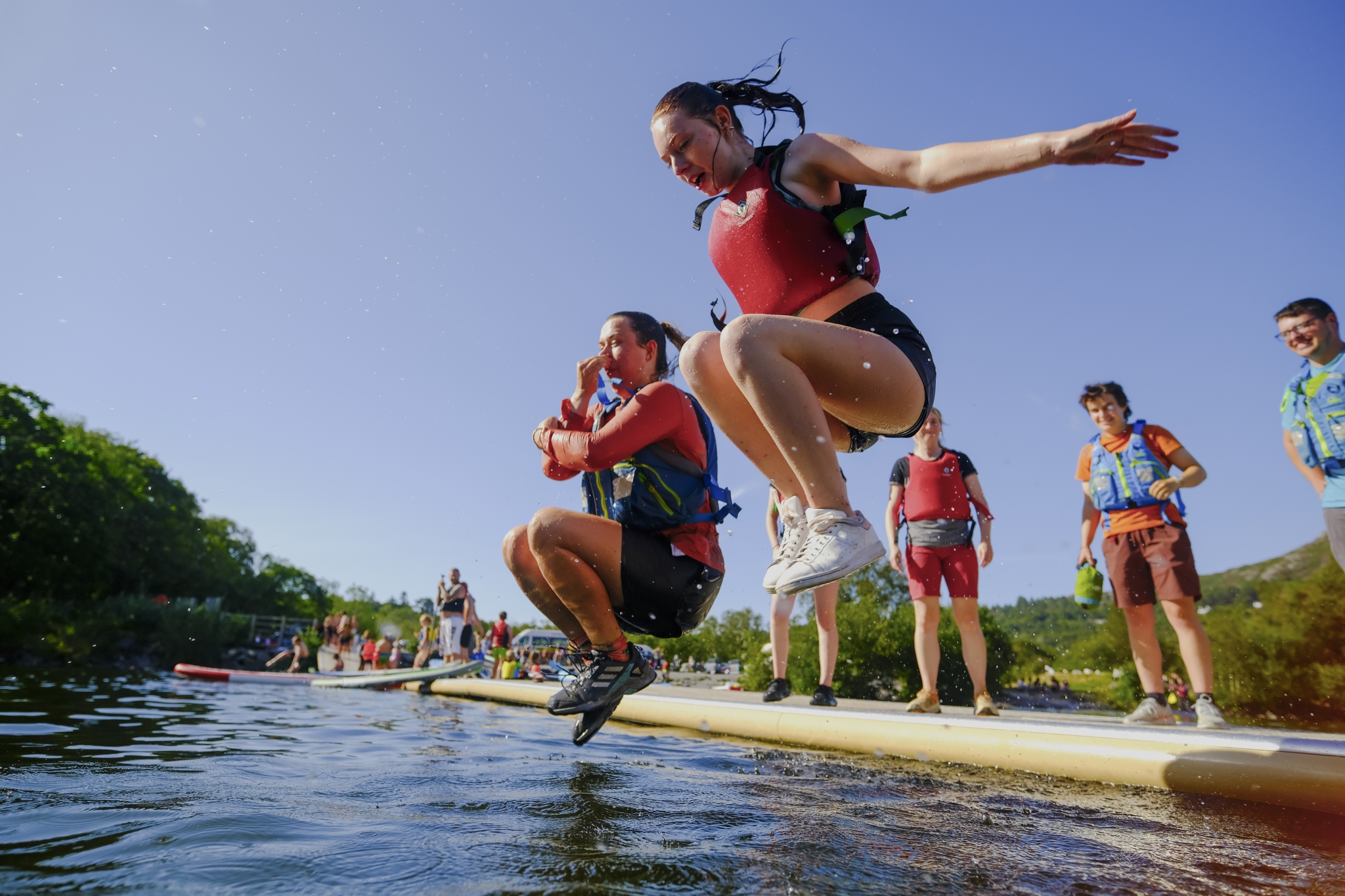 Girl jumping off a paddleboard into the water on a sunny day, with friends standing nearby under a bright blue sky—summer at Outward Bound.