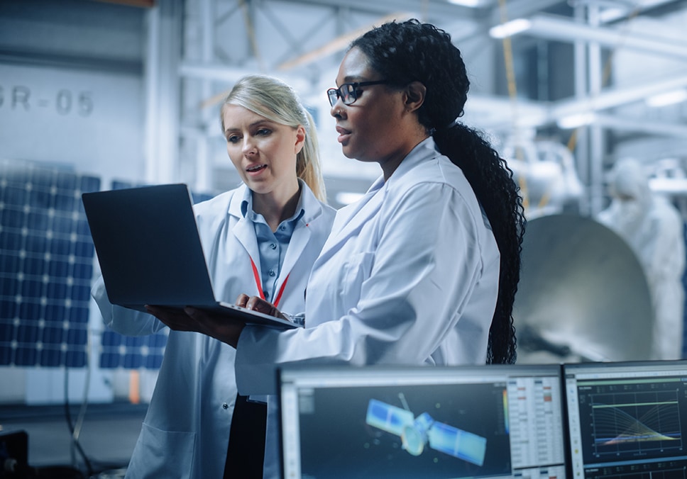 Research engineers looking at data on a laptop with a screen in front showing a satellite