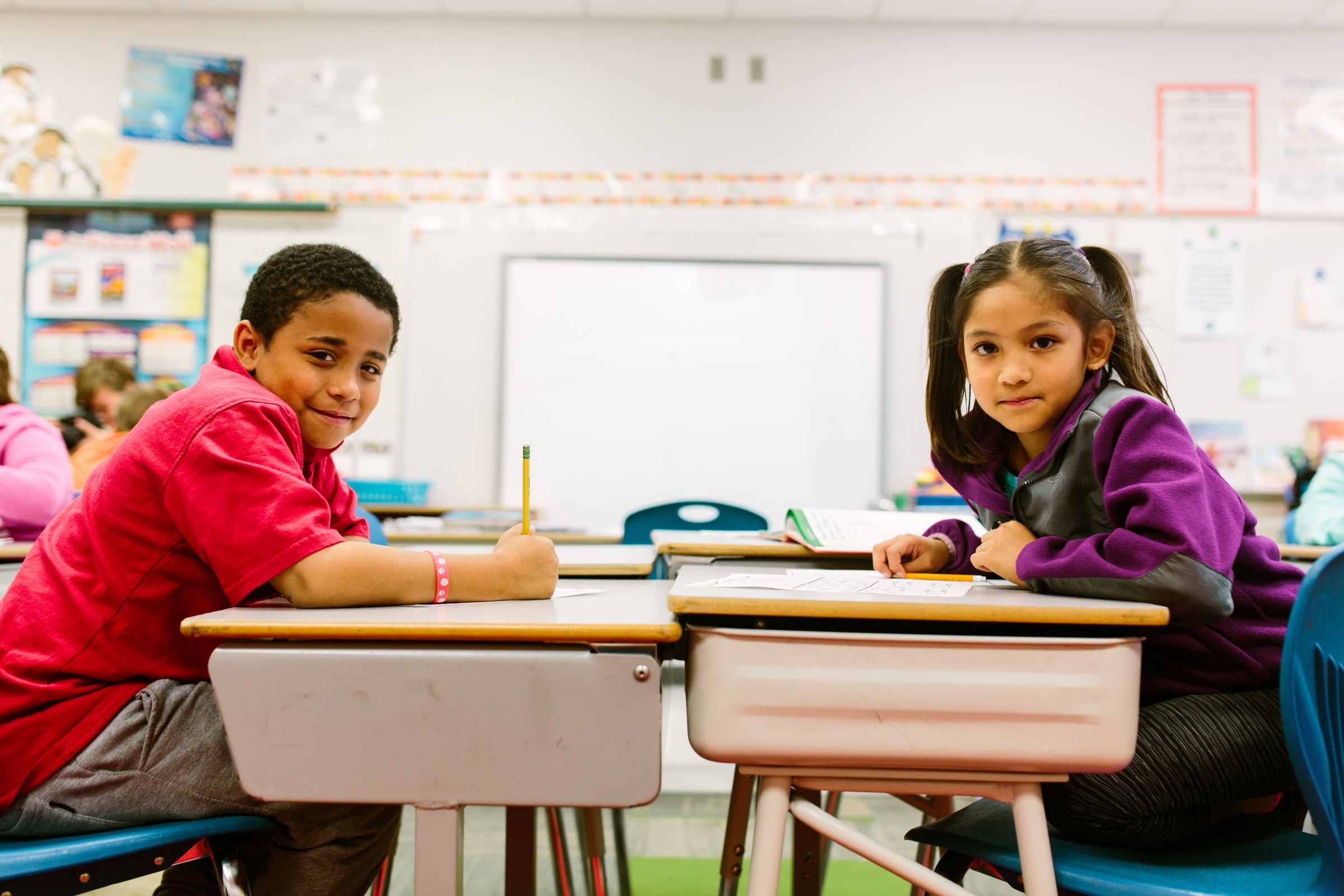 girl and boy at desks writing, smiling at camera