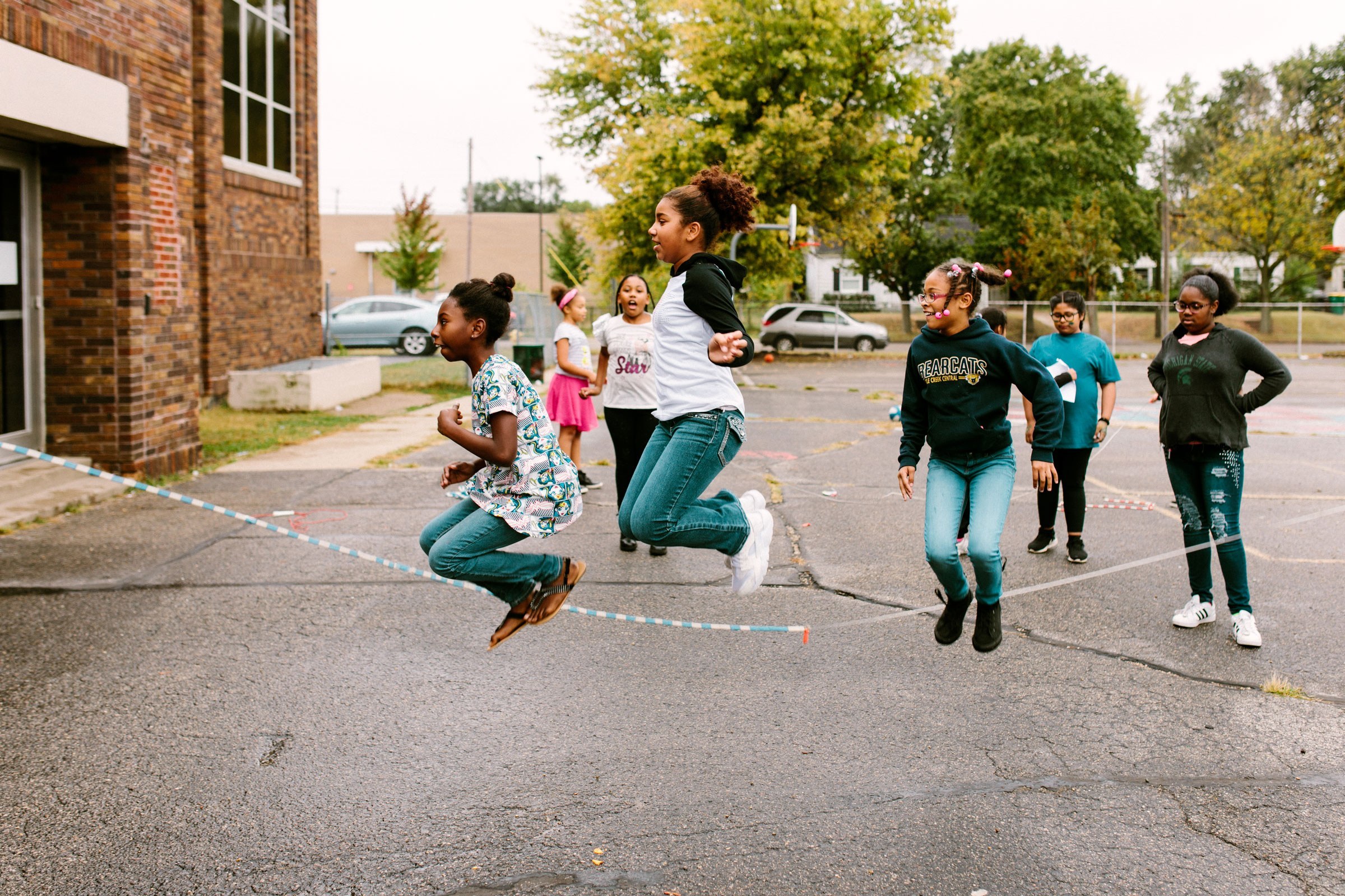 Three students jump rope in the yard while others cheer them on.