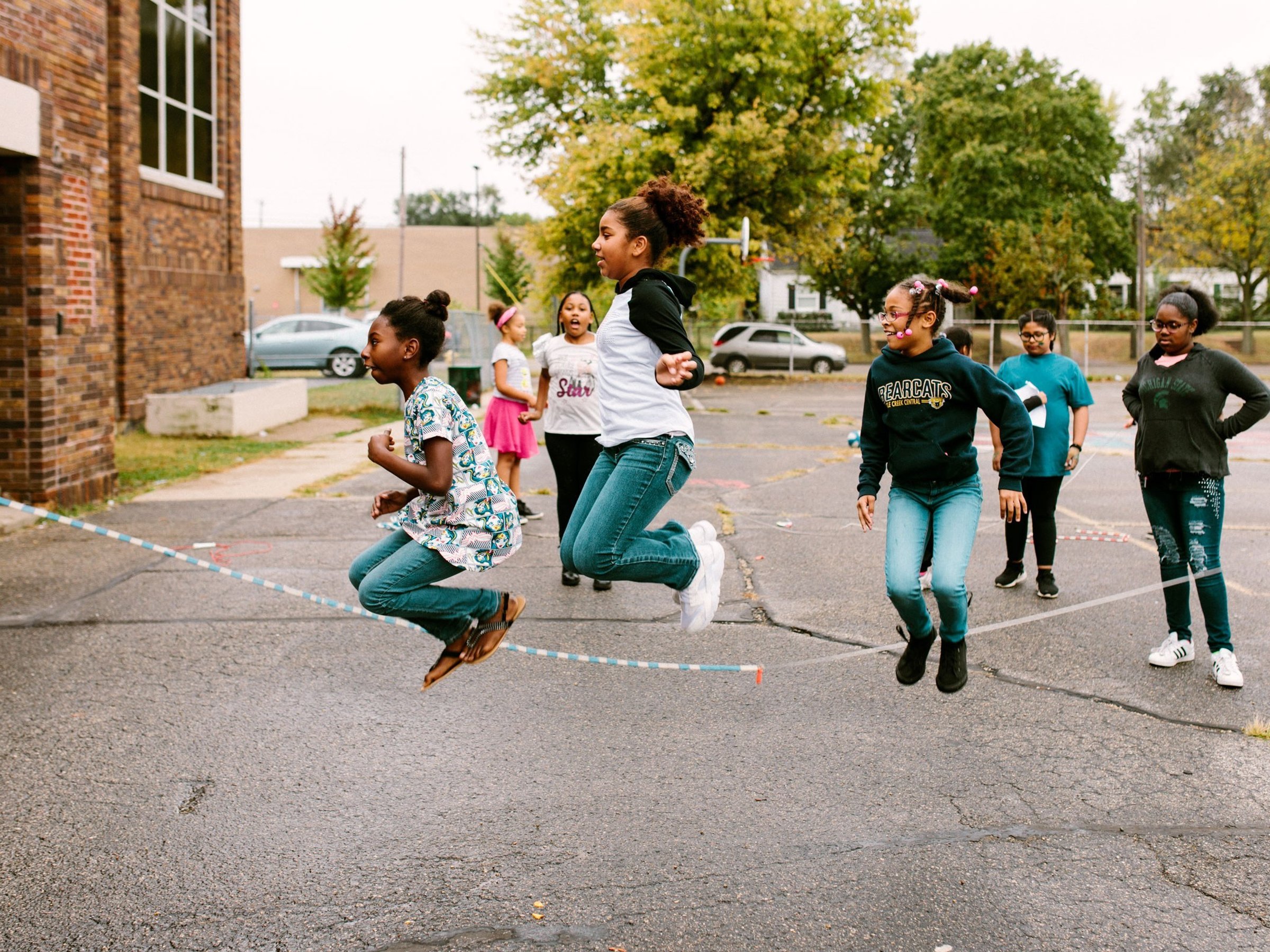 Three students jump rope in the yard while others cheer them on.