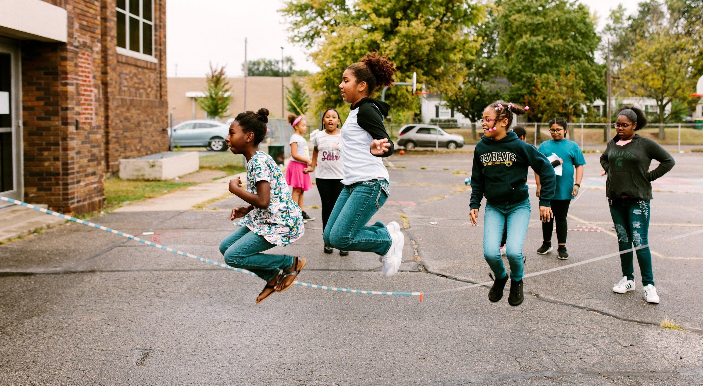Three students jump rope in the yard while others cheer them on.