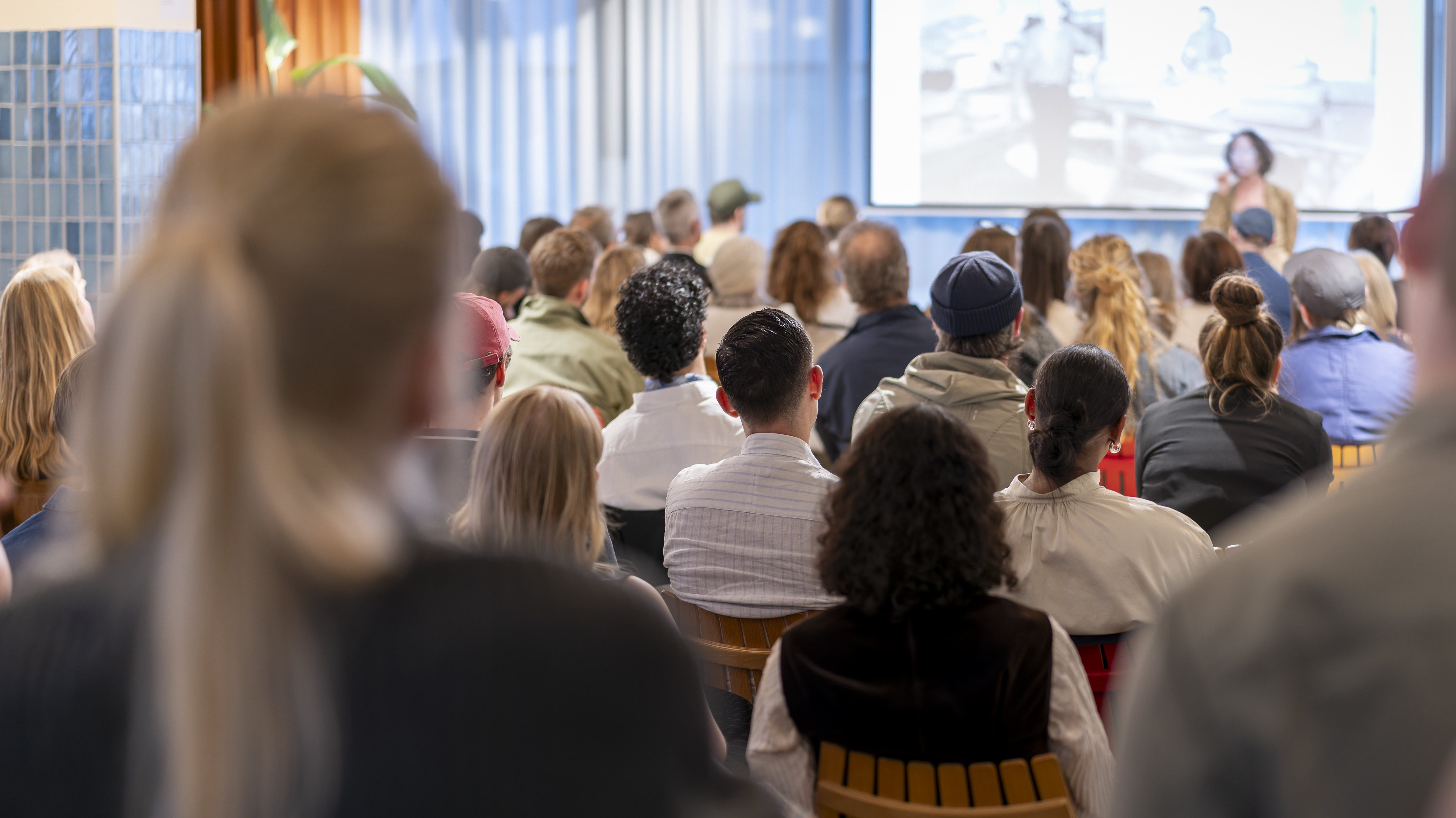 Tilhørere sett bakfra som følger med på en kvinne som presenterer foran en skjerm.