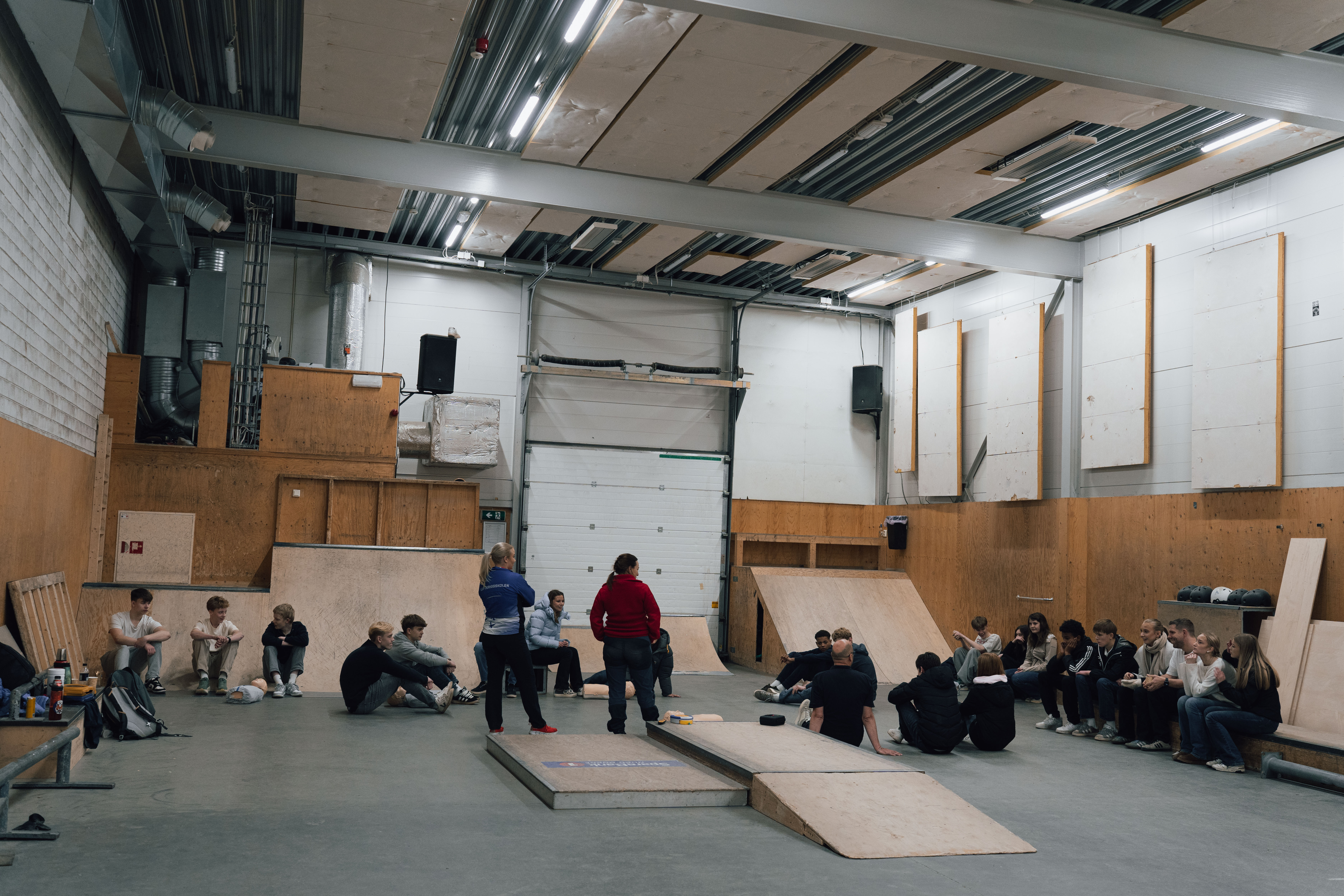 Foto av ungdommer som sitter spredt i en gymsal og lytter til to instruktører.