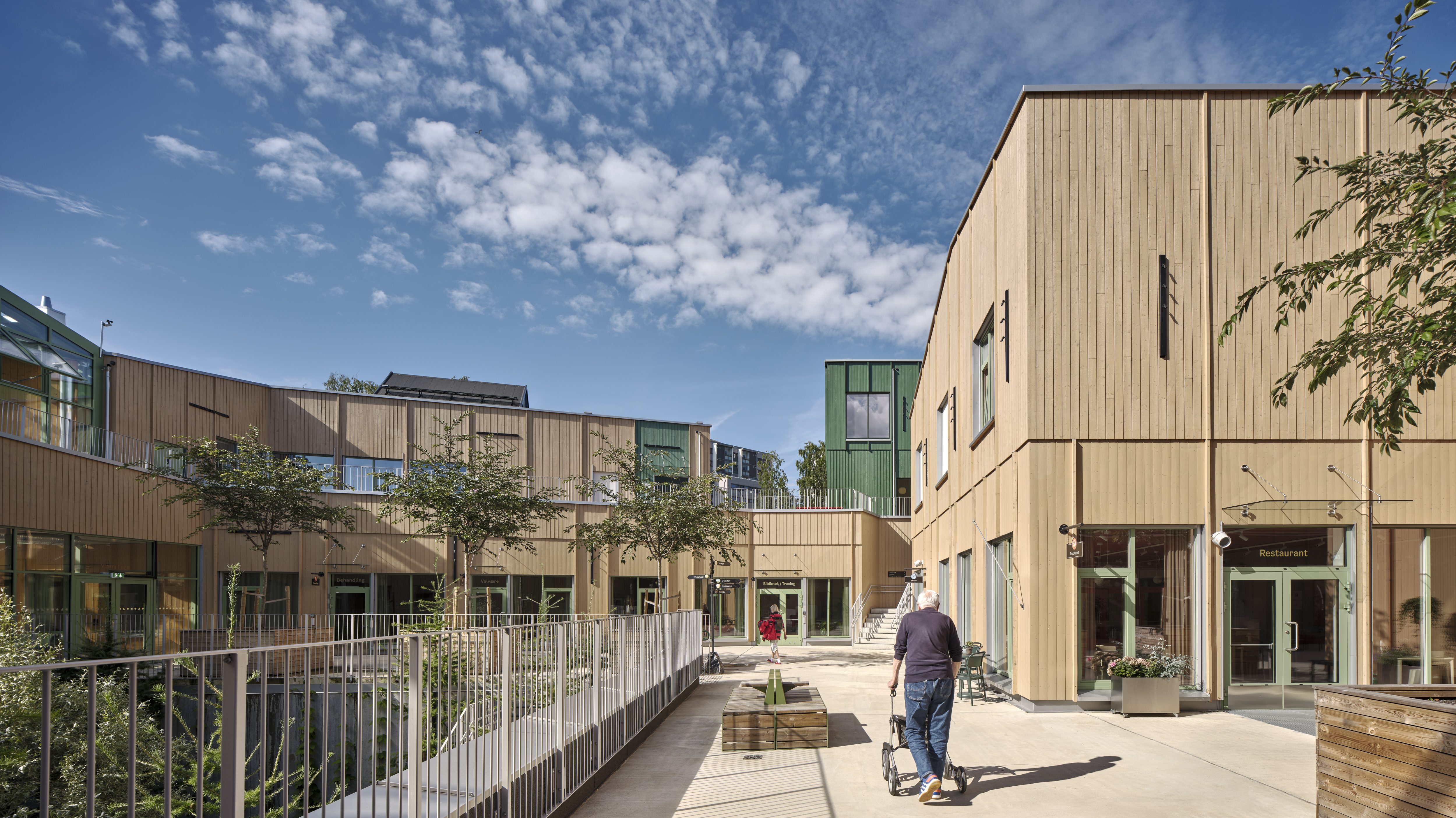 En mann med rullator går bortover mot lave bygninger med store vinduer.