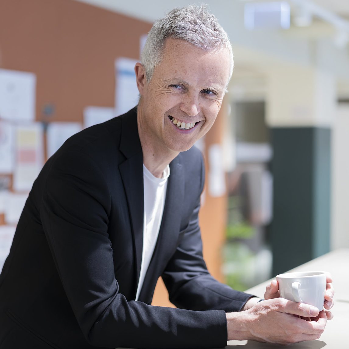 Foto av en smilende mann som lener seg over en hylle med en kaffekopp i hånda.