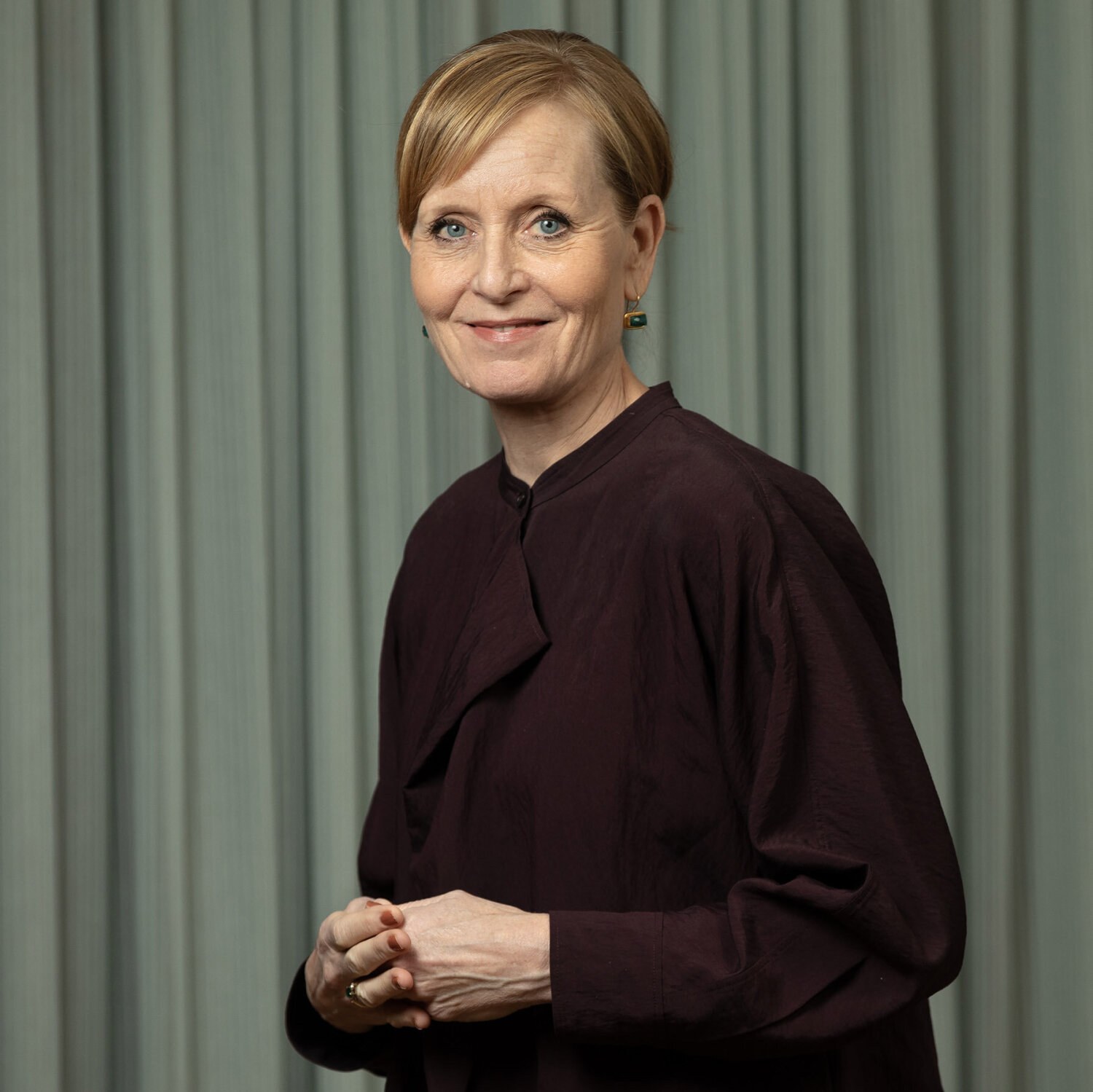 Foto av en smilende Benedicte med oppsatt blondt hår, burgunderrød bluse og øredobber med grønn stein.