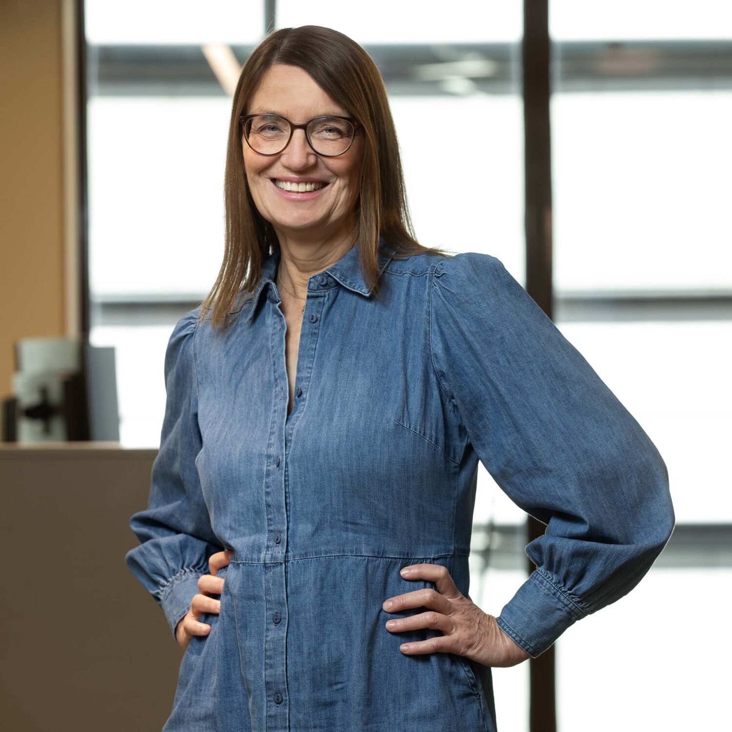 Foto av Ellen med briller og en kjole i denim, og smiler vennlig mot kamera.