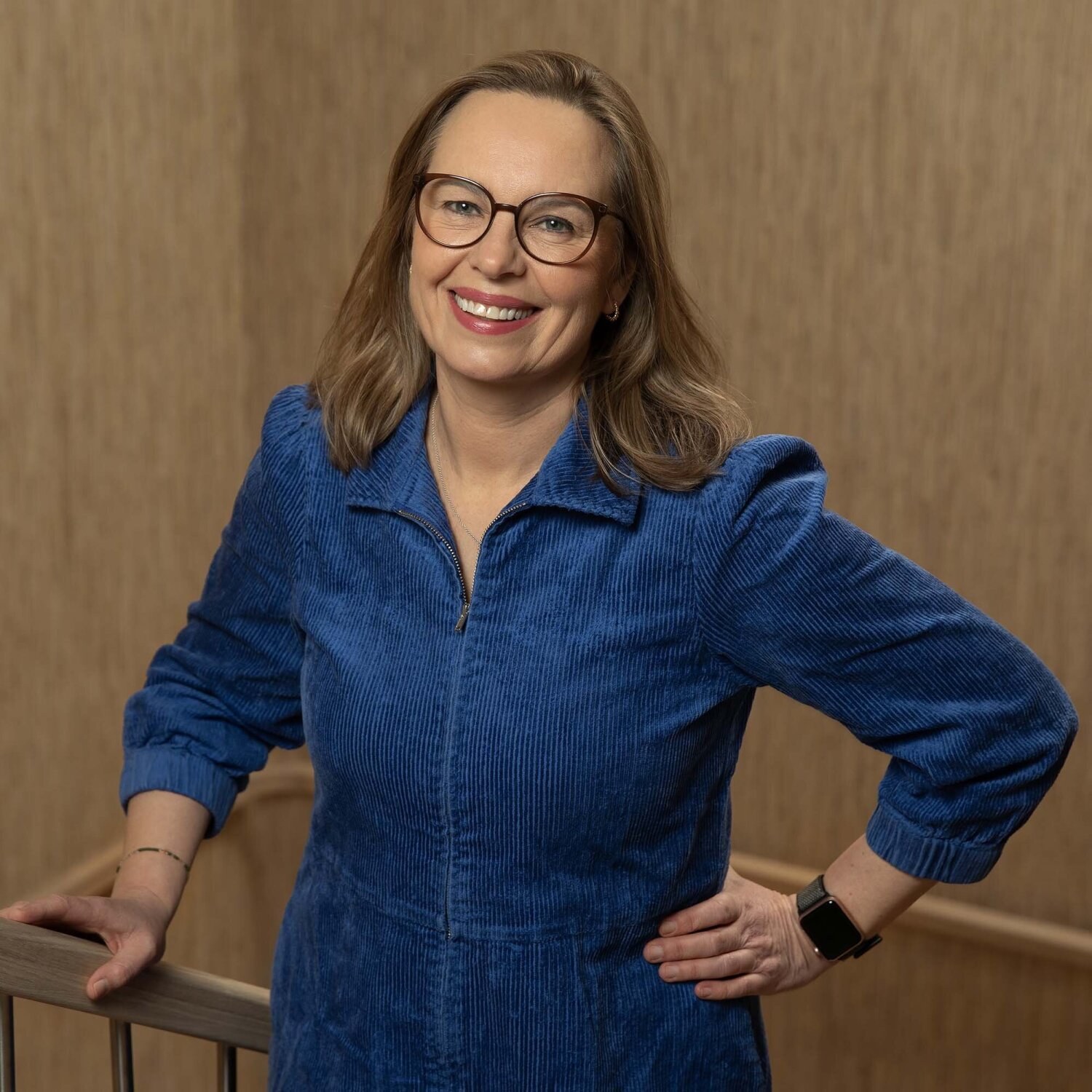 Foto av Eva som smiler mot fotografen i kornblå kjole og briller med mørk brun innfatning.