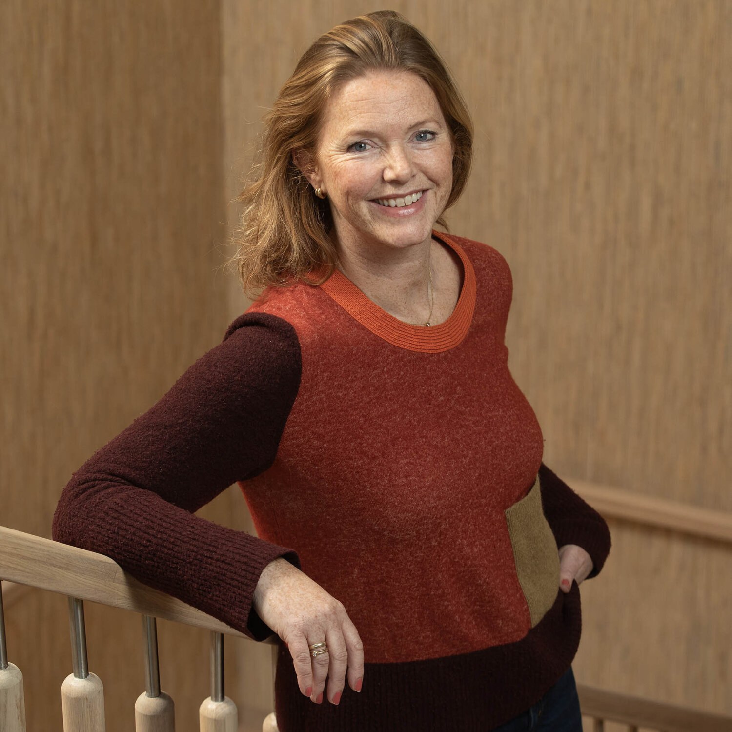 Foto av Hilde som står i trappen med et stort smil. Hun har rødlig, halvlangt hår gredd bakover og en topp i ulike rødbrune og beige fargetoner.