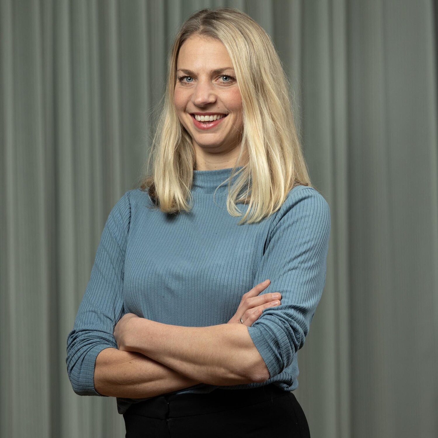 Foto av Ingeborg med armene i kors og et stort smil. Hun har halvlangt lyst hår og har på seg en dueblå genser.