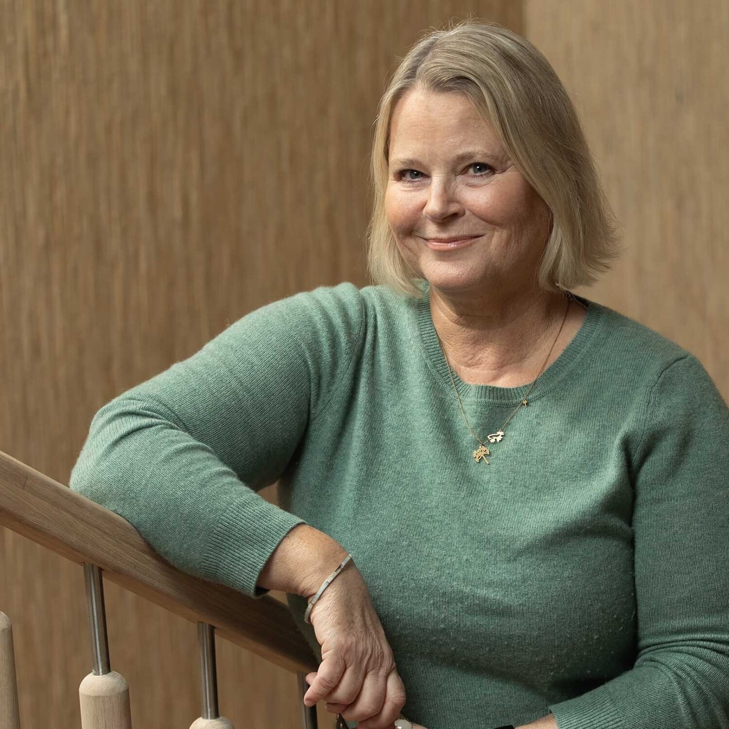 Foto av Jannicke som står lent opp mot trappegelenderet med et lunt smil. Hun har på seg en blågrønn genser og et tynt gullkjede med anheng.