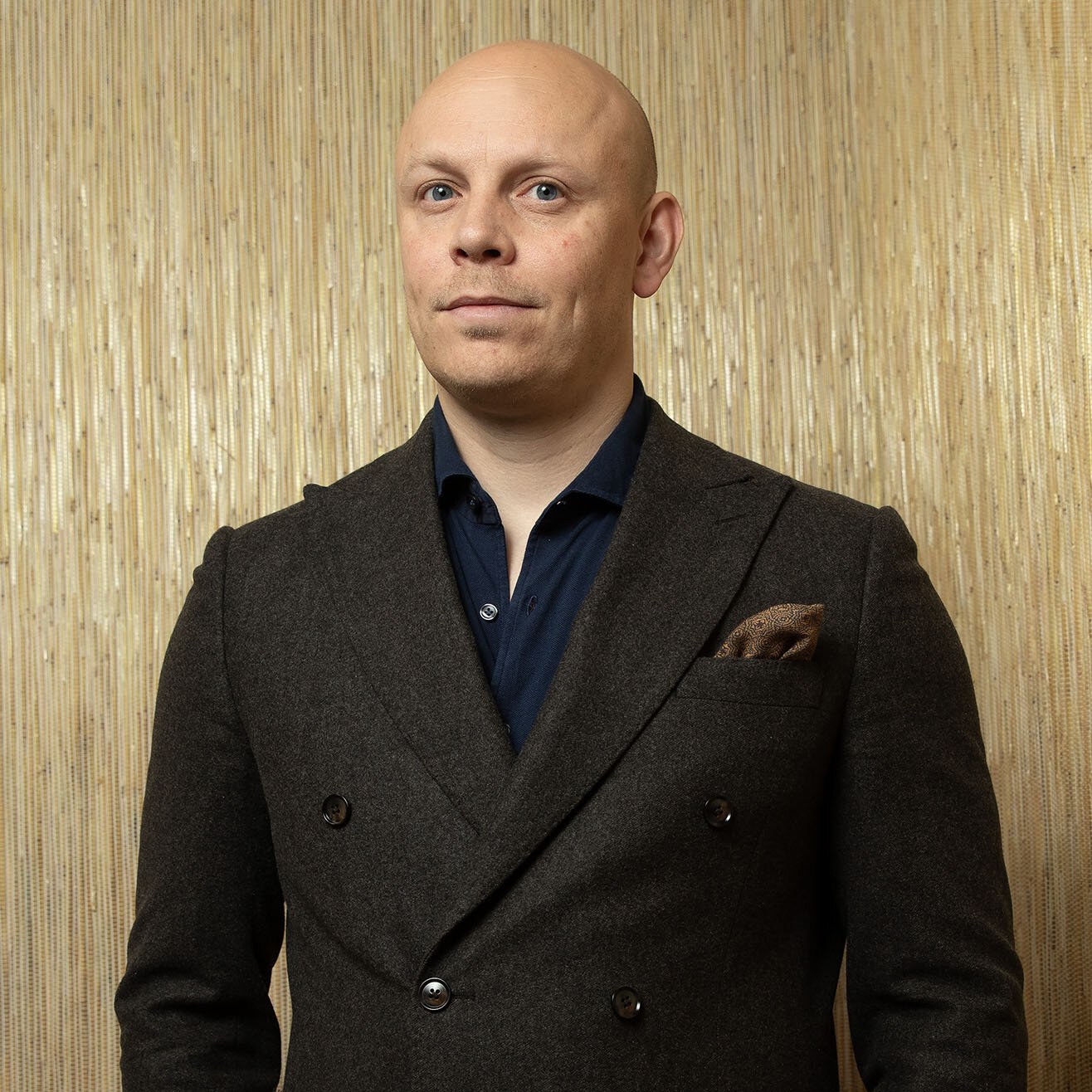 Foto aav Jørgen med et direkte, vennlig blikk og glattbarbert hode. Han har på seg blå skjorte og brun blazer med et tørkle stikkende opp fra brystlommen.