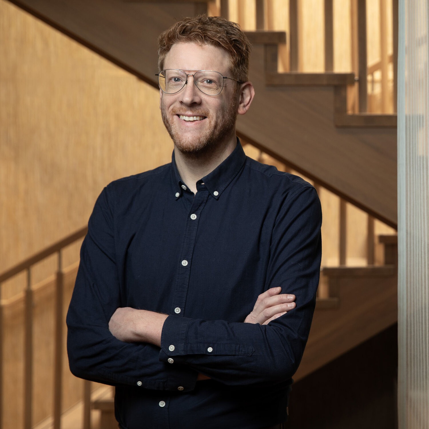 Foto av Med kort, rødt hår og briller står Martin med et vennlig smil, Han har på seg en marineblå skjorte og står med armene i kors foran en trapp.