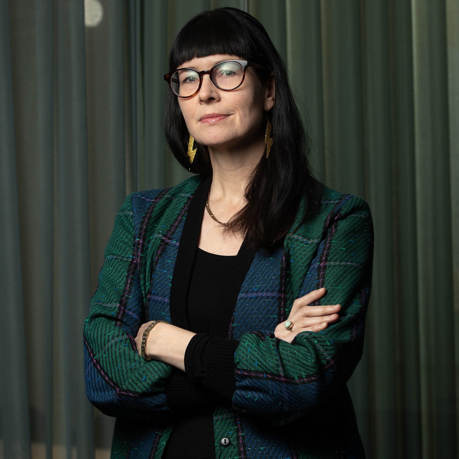 Foto av Matti med et lite smil om munnen, sort hår, pannelugg og briller. Hun bærer en skotskrutet jakke over en sort topp.