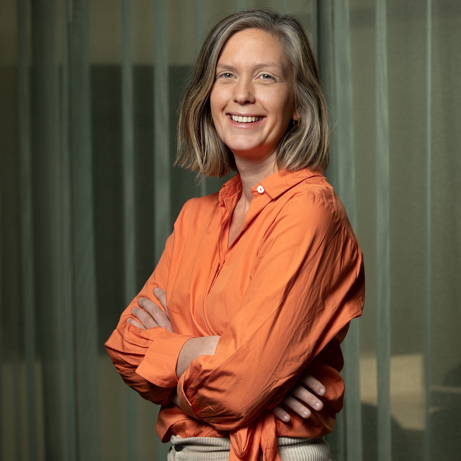 Foto av en smilende Siri med halvlangt blondt hår og oransje bluse.