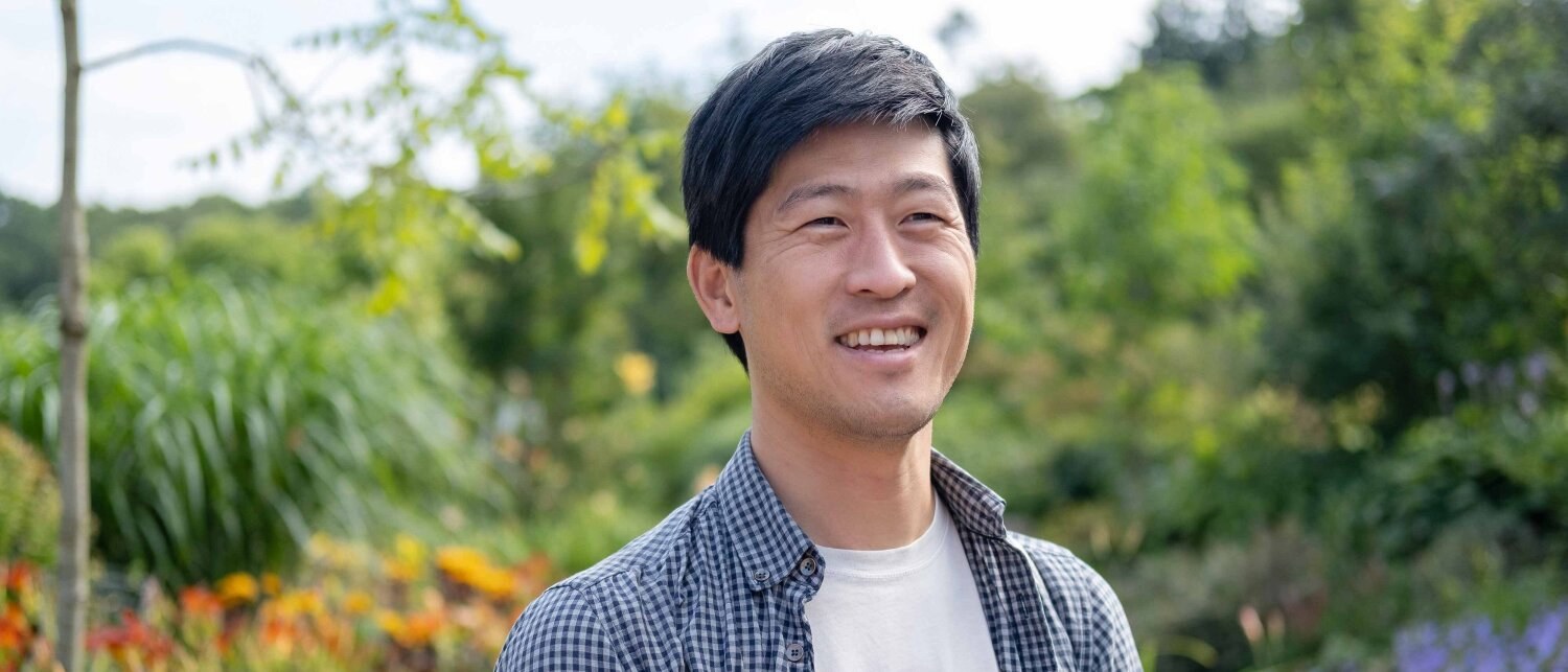 Foto av Svein smilende mot kamera mens han står ute i grønne omgivleser.