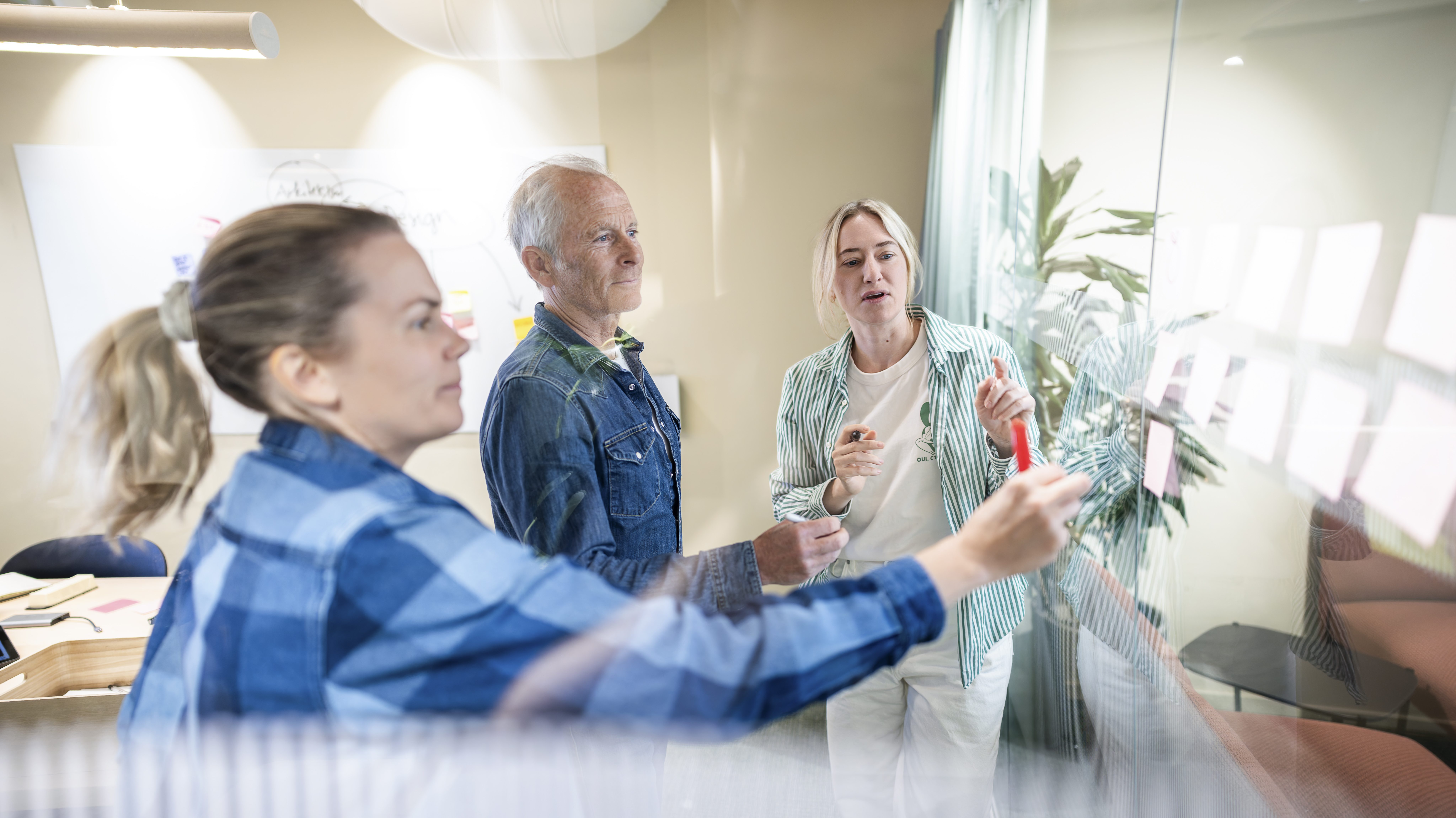 Tre personer står ved en glassvegg og diskuterer mens de plasserer post-it-lapper.