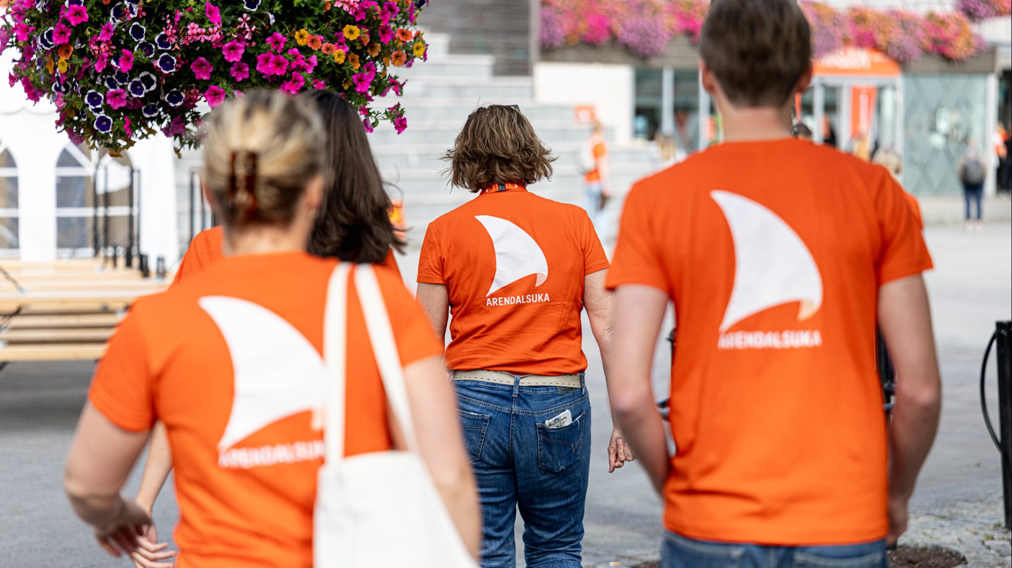 Tre personer med ryggen til går bortover en gate med blomsteroppsatser. De har på seg oransje t-skjorter med logo fra Arendalsuka.