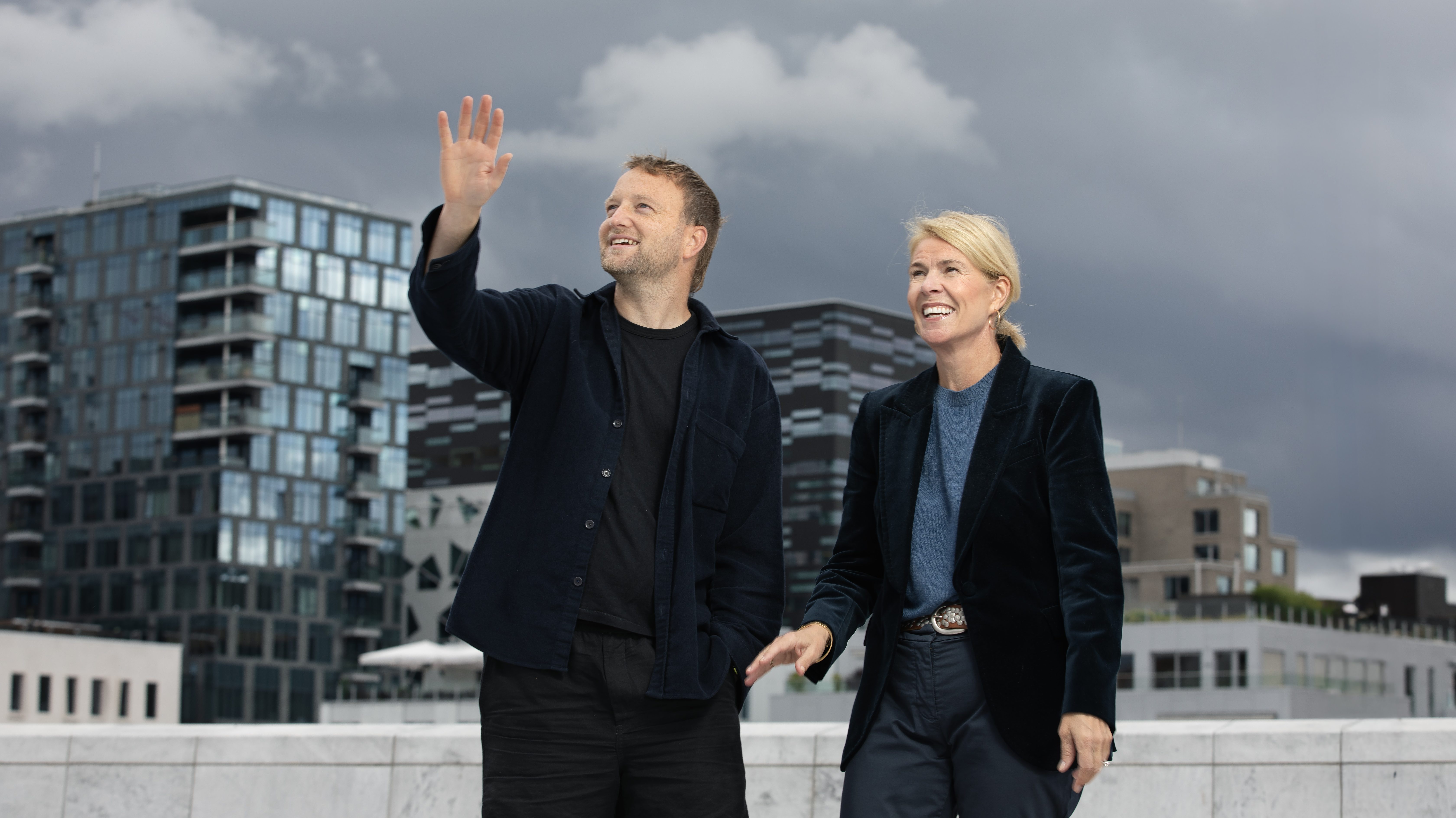Ask Helseth står med armen hevet foran forretningsbygg i Bjørvika i Oslo. Ved siden av ham står Anne Bull. Begge er kledd i sort.