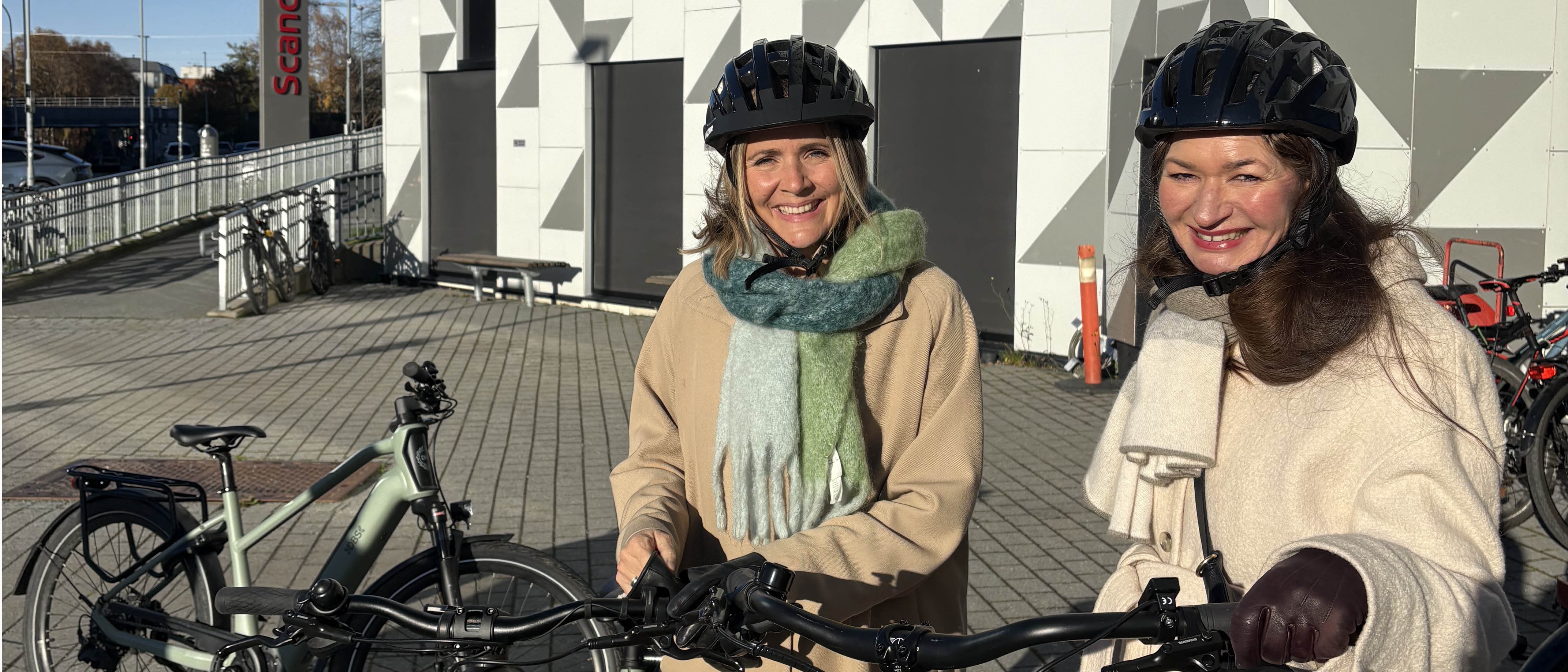 To blide kvinner med svart sykkelhjem står foran en bygning og holder hver sin sykkel.