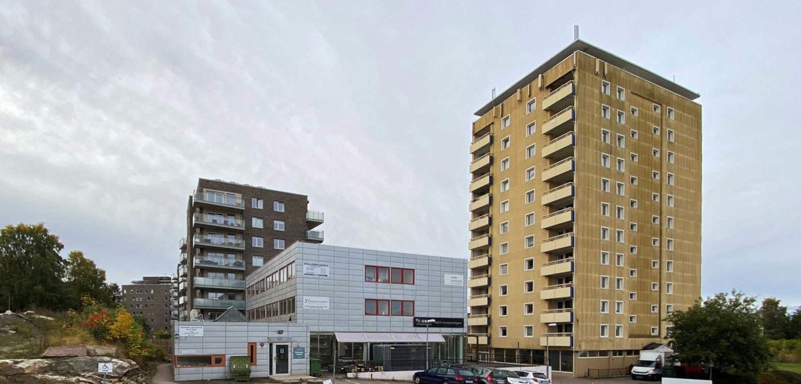 Til høyre en gul boligblokk med mange etasjer, typisk for boligbygg fra 1960-70-tallet. Midt i bildet en lavere, mer industriell bygning med fasade av lyse metallplater. I bakgrunnen, flere boligblokker.