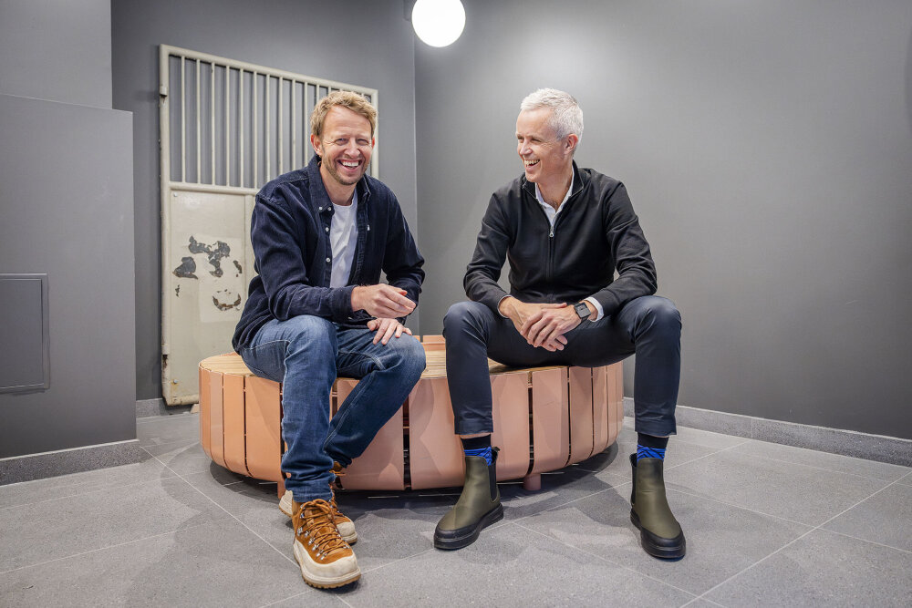 Foto av to menn som sitter smilende på en benk.