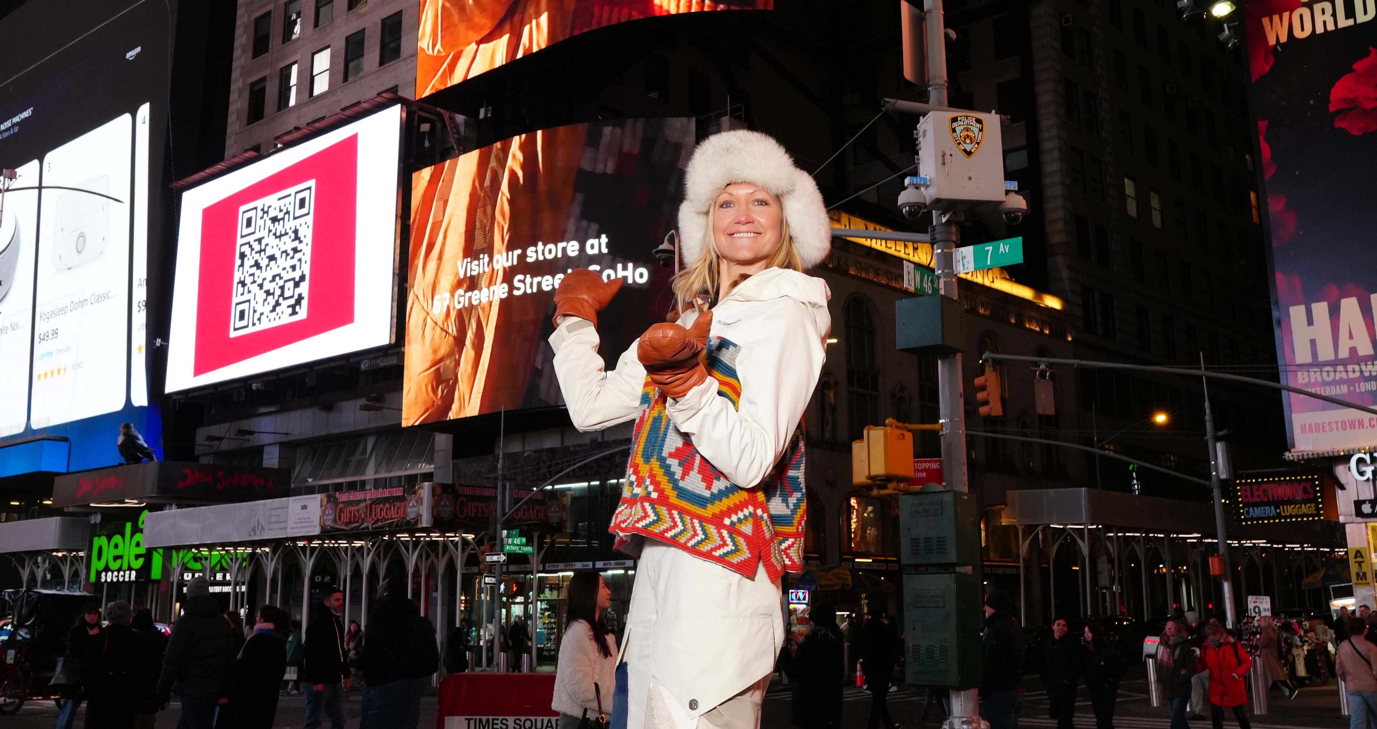 En blid dame kledd i friluftsklær og en stor pelslue poserer foran reklameplakater på Times Square i New York.
