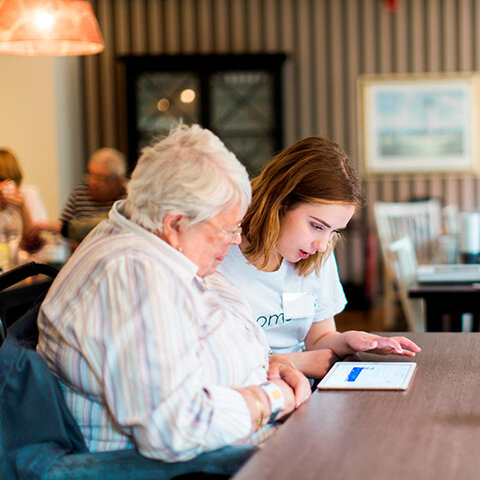 Foto av en yngre jente som hjelper en eldre dame med et nettbrett.