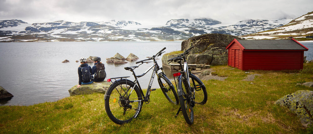 Foto av to sykler som står parkert ved et fjellvann med spektakulær utsikt til snøkledde topper.