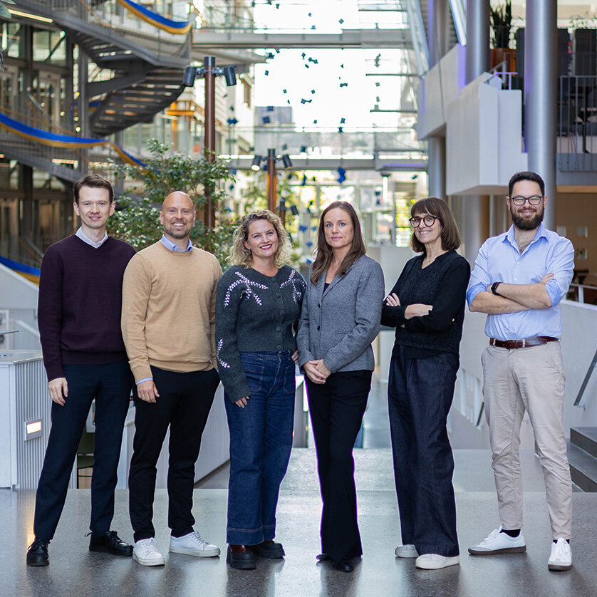 Foto av seks mennesker som står samlet, smilende mot kamera inne på BI Oslo sin inngangshall.