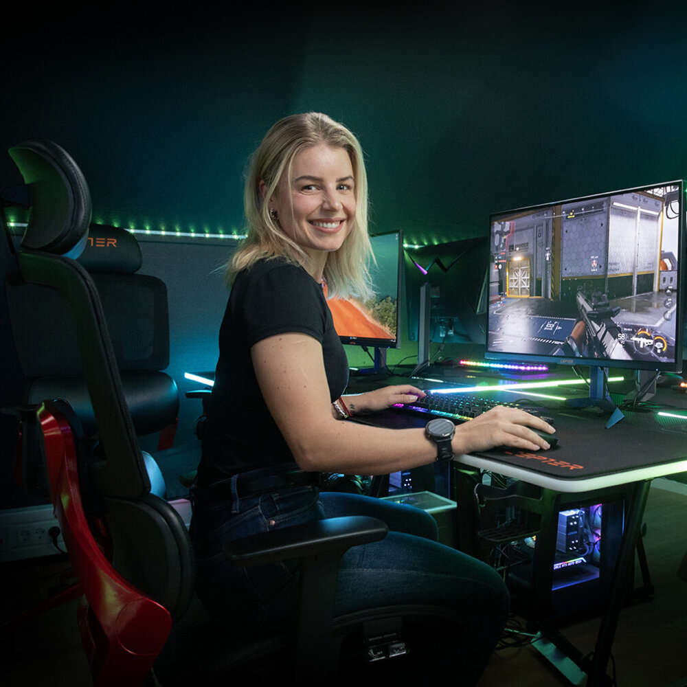 Foto av en dame som sitter foran en opplyst PC med spill på, og smiler mot kamera.