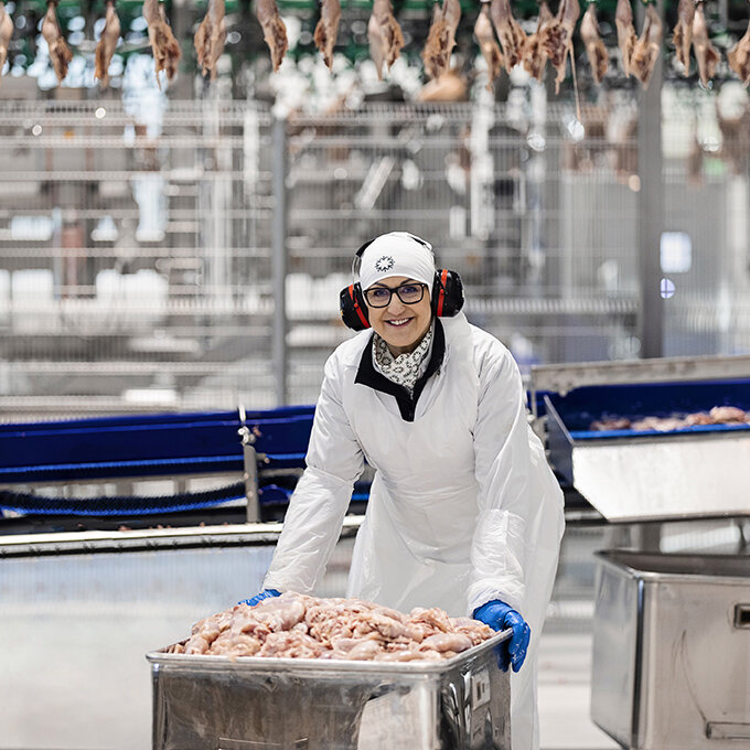 Foto av en smilende arbeider i hvit beskyttelsesdrakt, hørselsvern og vernebriller i en matproduksjonshall for kylling.