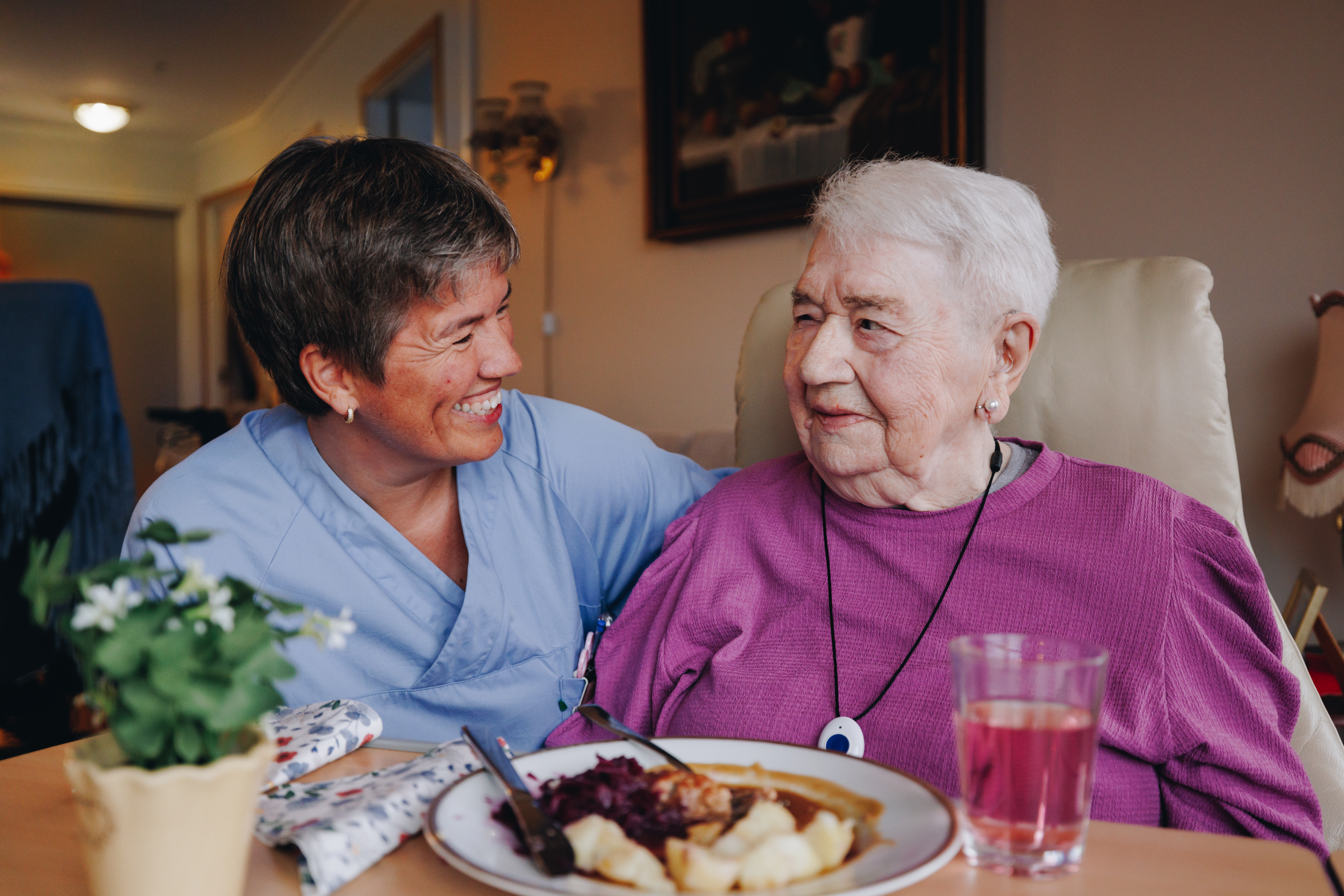En omsorgsarbeider i blå overdel ser smilende bort på en eldre kvinne med lilla genser. De sitter ved et lite bord der det står en middagstallerken og et glass rød saft.