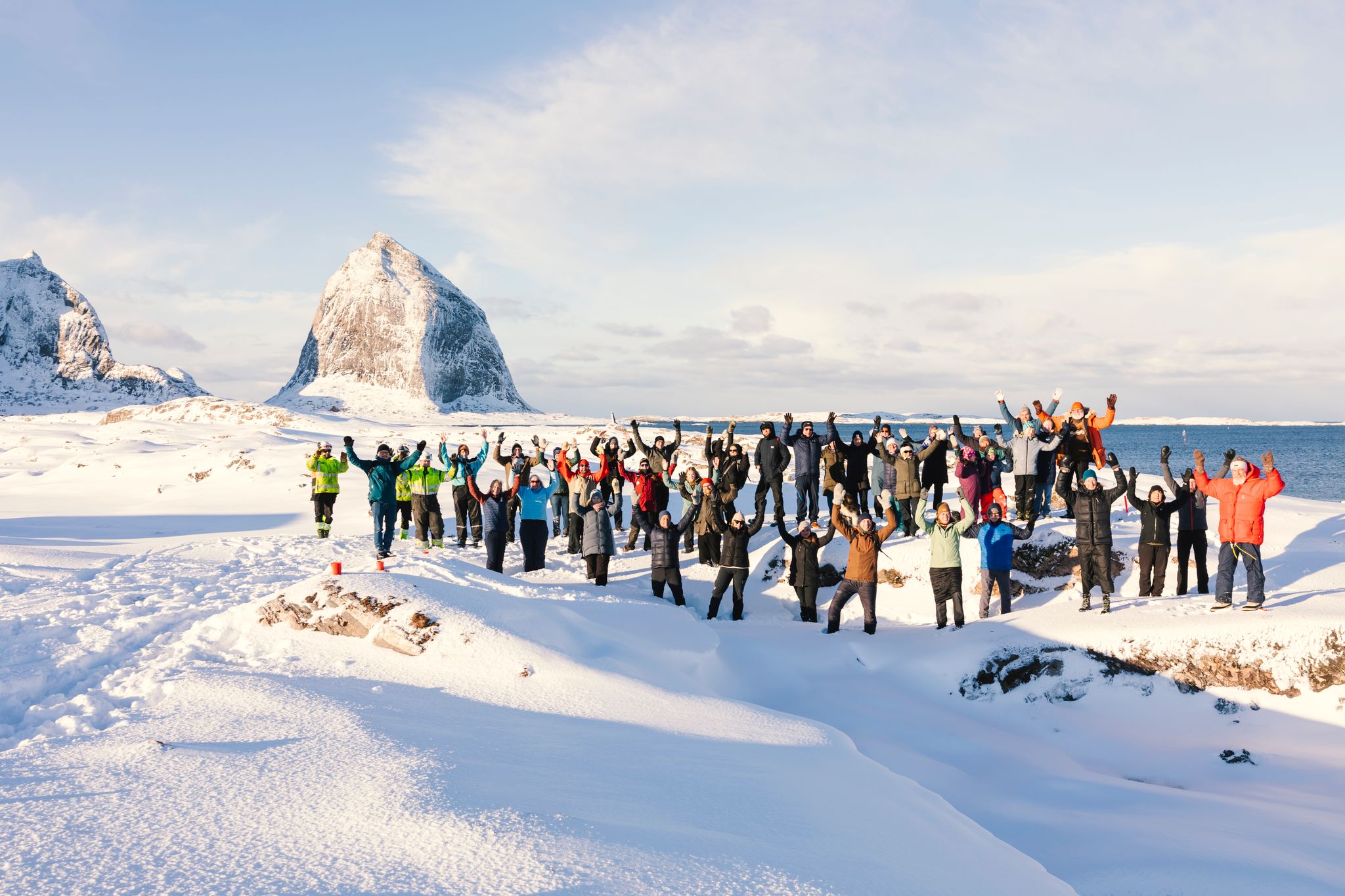Mange mennesker står med armene i været i et snøkledd vinterlandskap foran en stor fjelltopp.