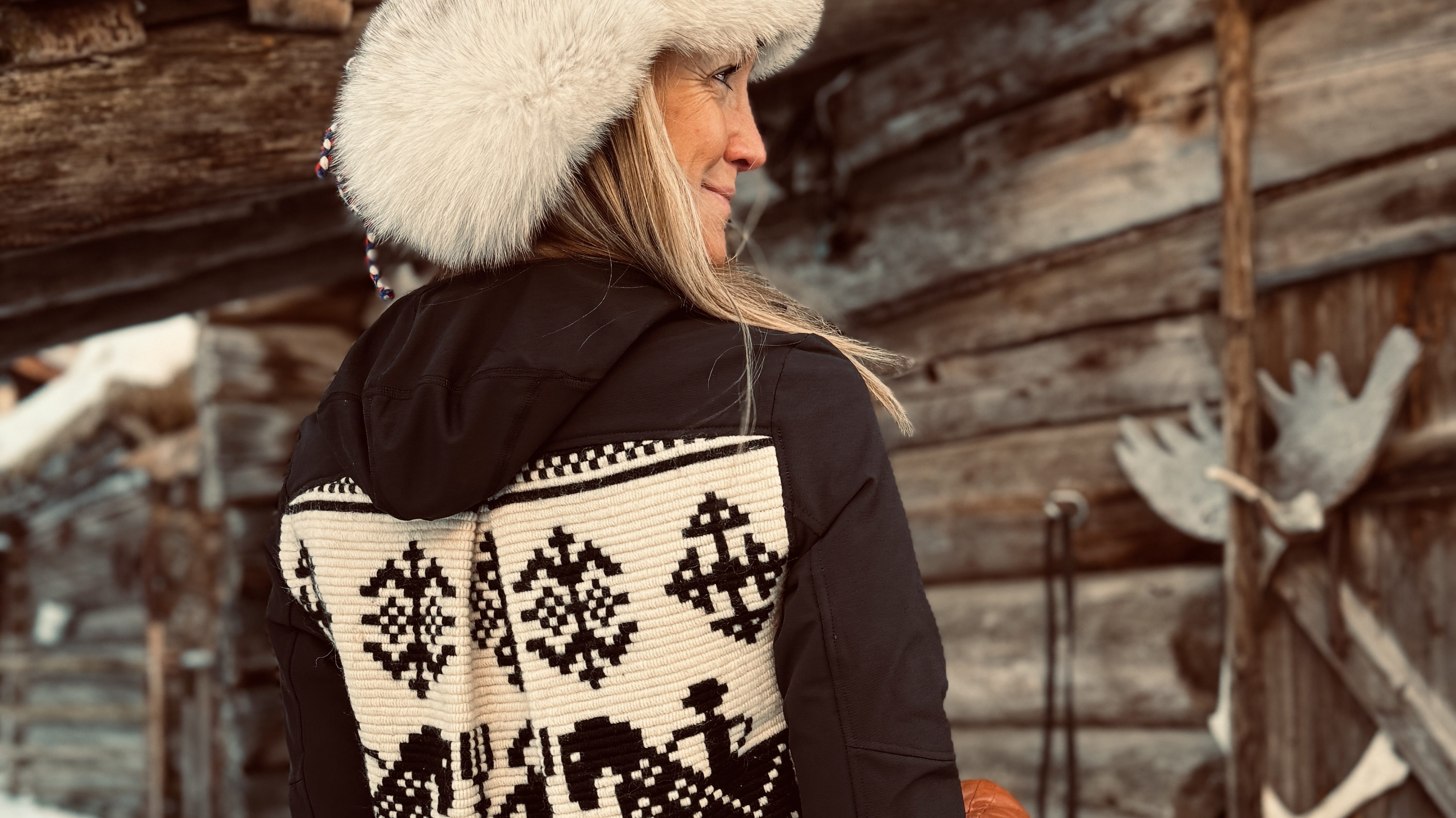 En dame sett bakfra med stor pelslue og en jakke med elementer fra tradisjonelle norske mønstre står foran en slitt tømmervegg.