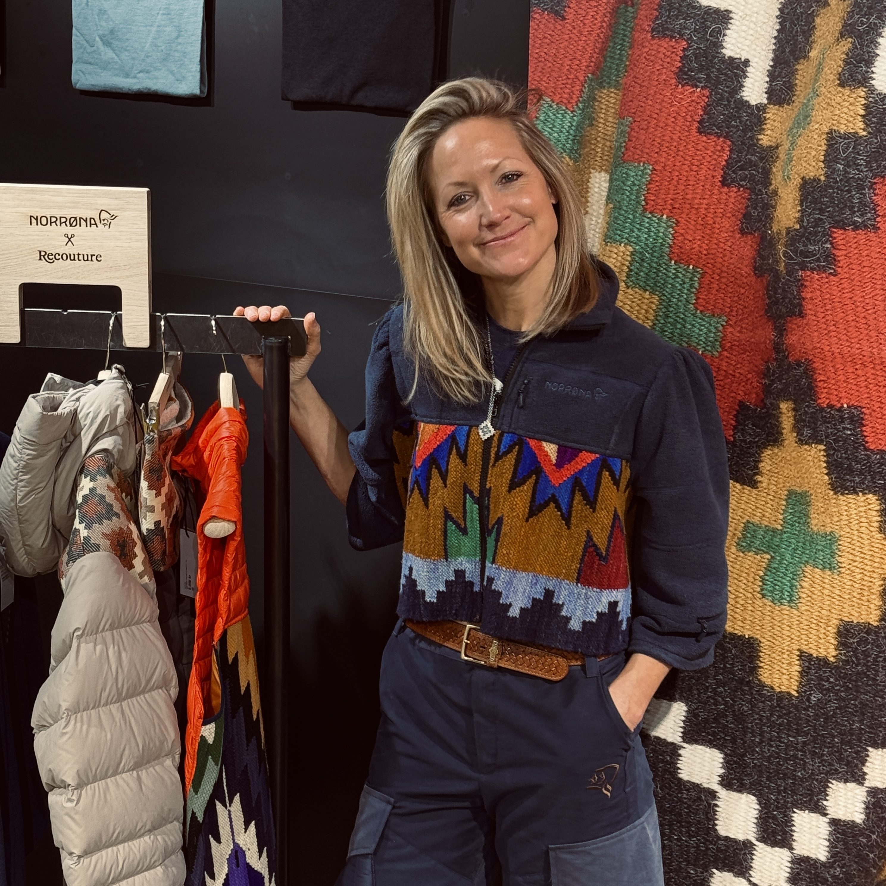 En smilende dame med lyst hår står foran et veggteppe med tradisjonelle norske mønstre og et stativ med friluftsklær.