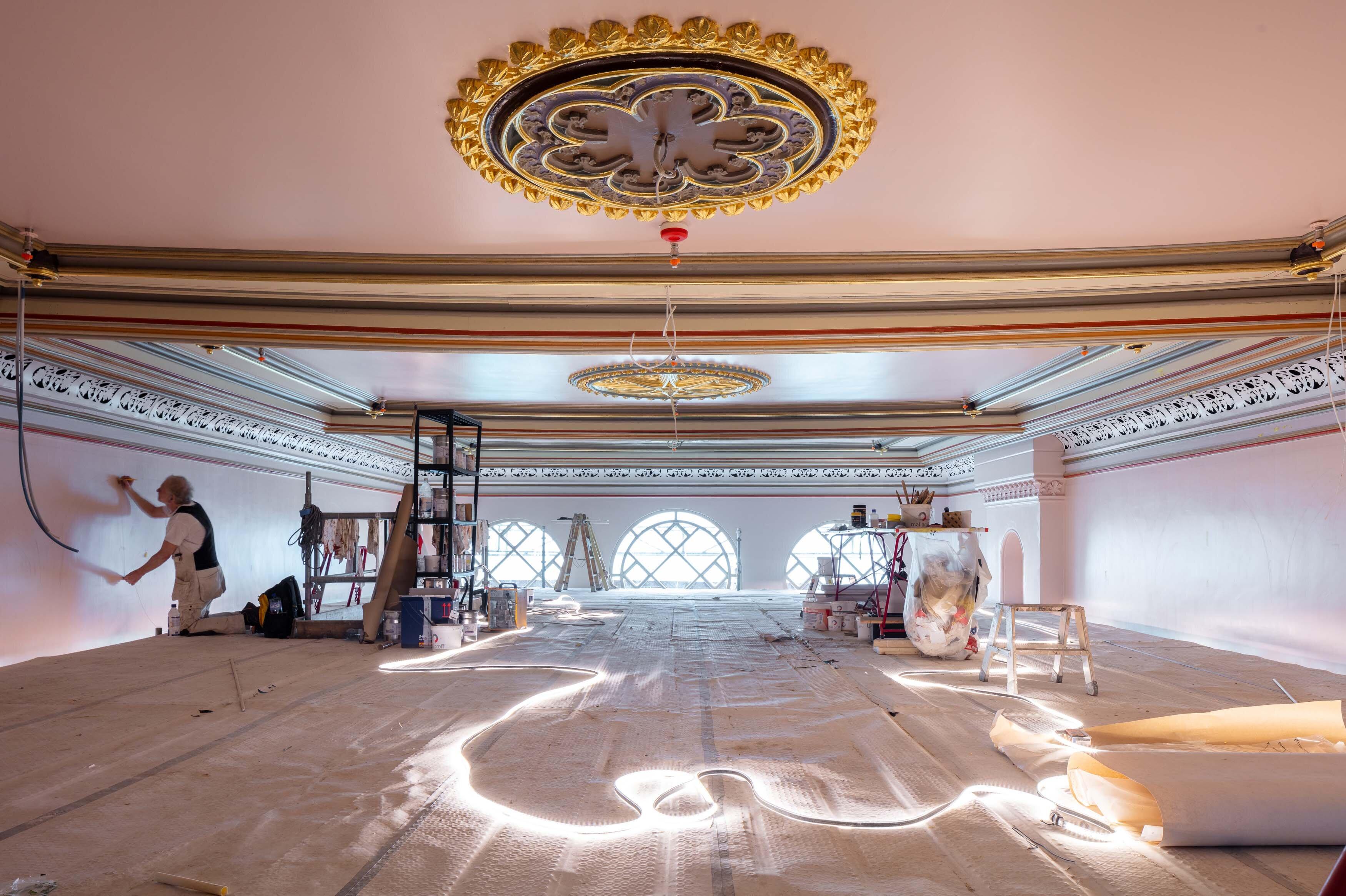 Interiørbilde fra pågående restaureringsarbeid. En håndverker arbeider på et stillas plassert helt oppunder taket hvor det ses nymalte rosetter og ornamentering.