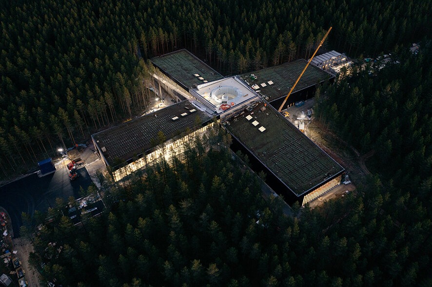 Oversiktsbilde av fabrikk i en furuskog. Fabrikken ligger som en pluss, men skogen tett på. Det er mørkt, men lyset fra vinduene utgjør et lysende omriss rundt fabrikken.