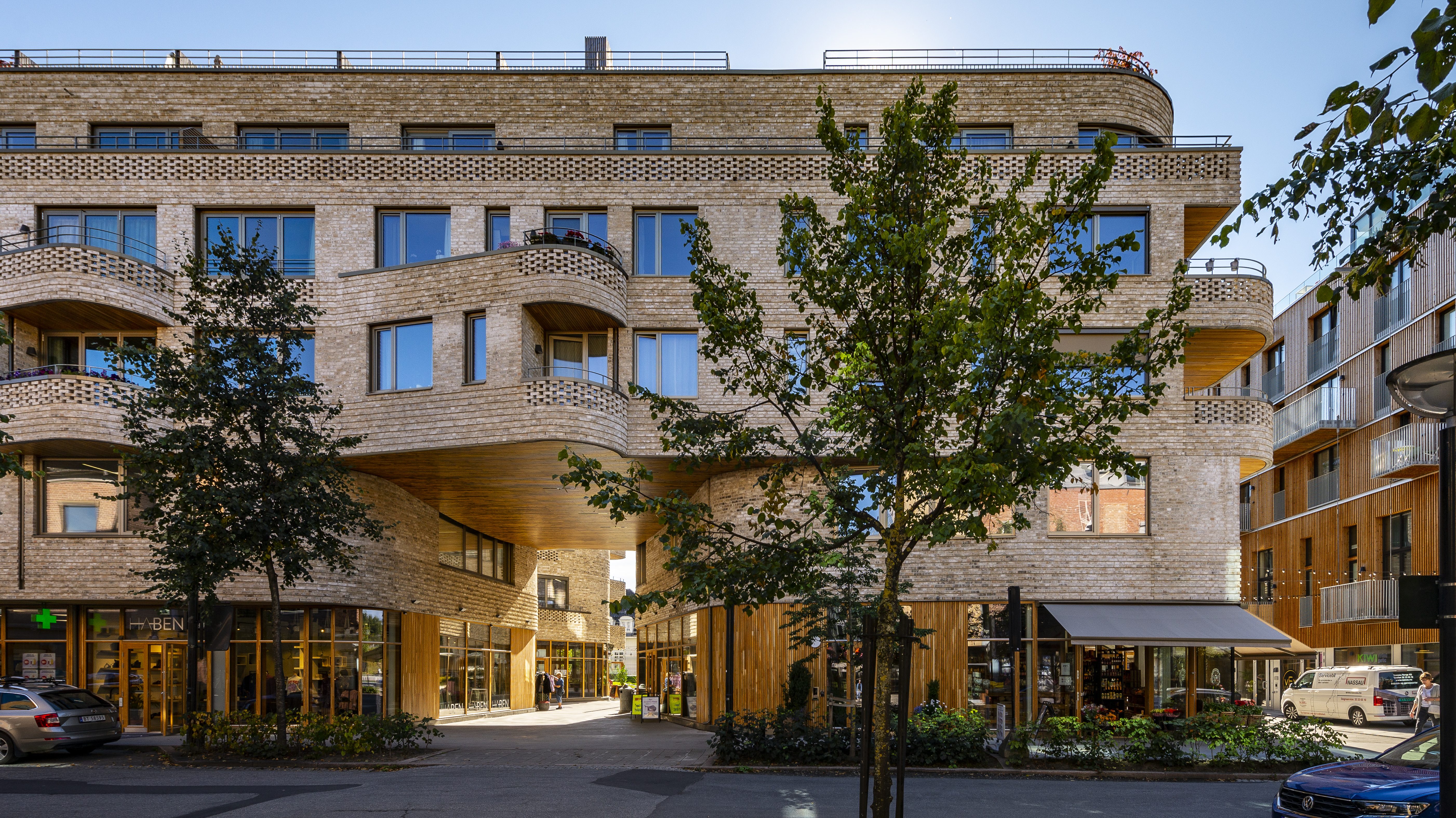 Moderne bolig- og næringsbygg i lys tegl med avrundede balkonger og detaljer i tre. Bygget har flere etasjer med store vinduer og butikker i første etasje. Et åpent passasjeområde går gjennom bygget i bakkeplan. Trær langs fortauet rammer inn fasaden, og parkerte biler står i gaten foran. Det er solfylt vær og blå himmel.