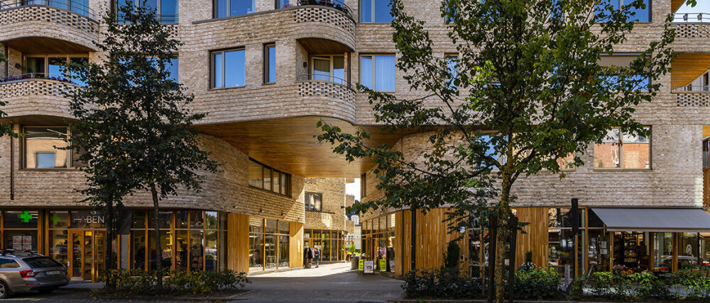 Lyst mursteinsbygg med to trær foran og åpen passasje gjennom bygget. Foto.