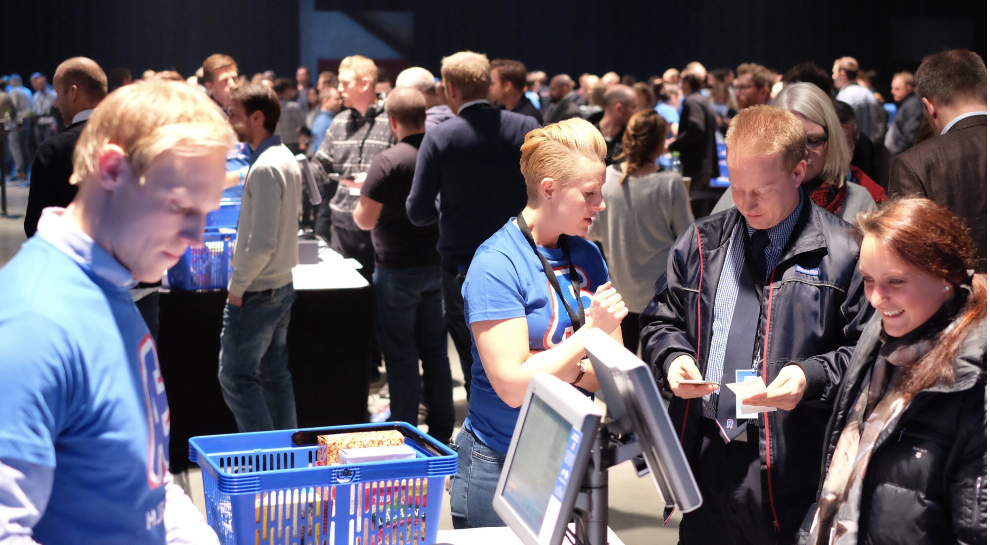 En folkemasse samlet i et messelokale. To fremme i bildet er ikledd blå "Ækspert-t-skjorter". De ser ut til å drive opplæring av deltakere rundt en fiktiv Rema 1000-kasse.